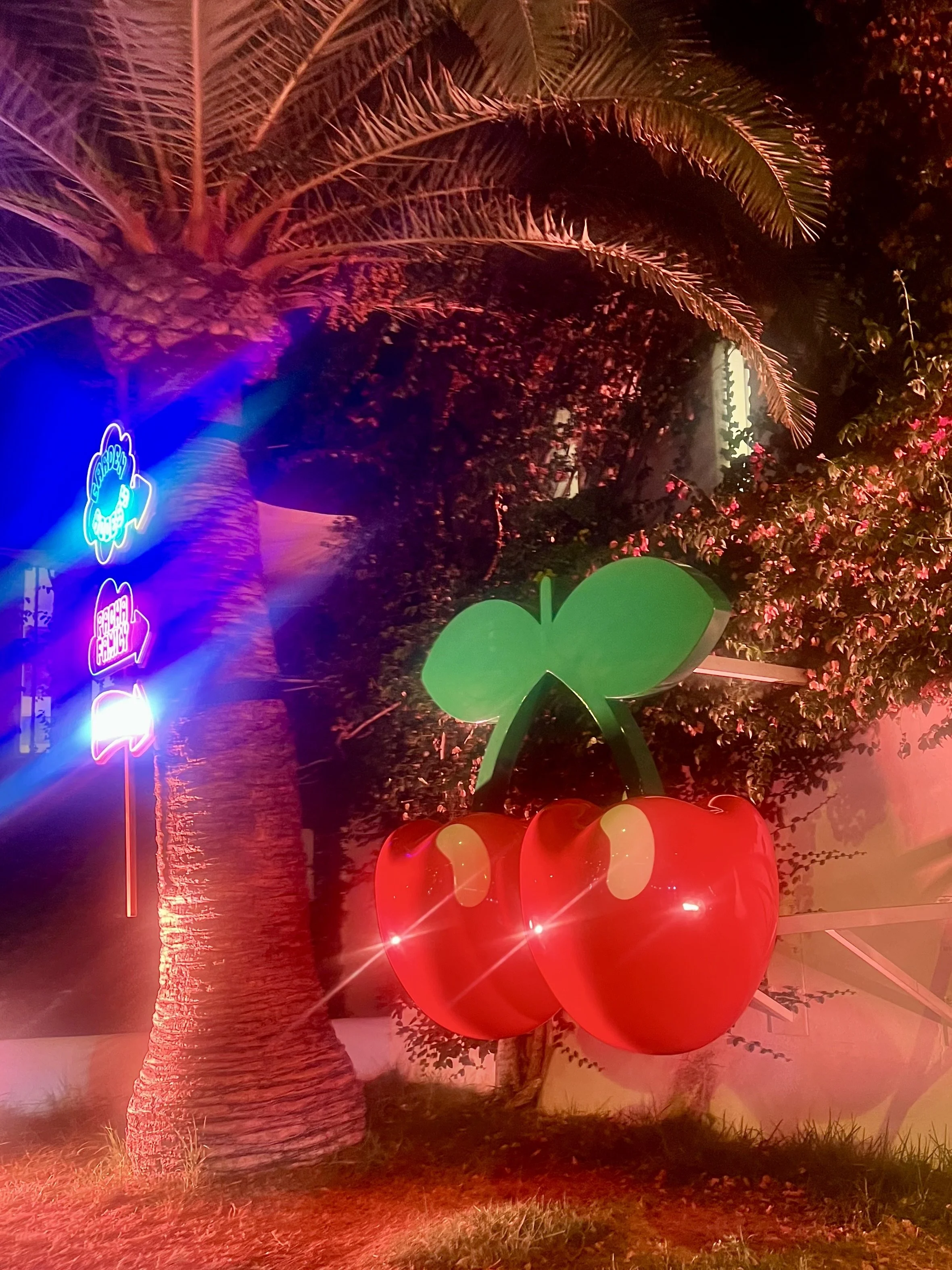 Night scene with illuminated palm trees, large red heart-shaped balloons, and colorful neon signs.