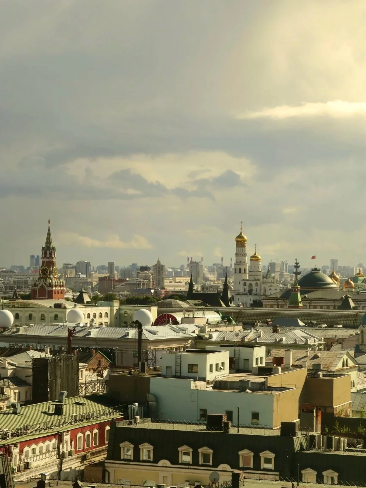 A city skyline featuring a mix of historic and modern buildings, including a church with golden domes and a clock tower, under a cloudy sky.