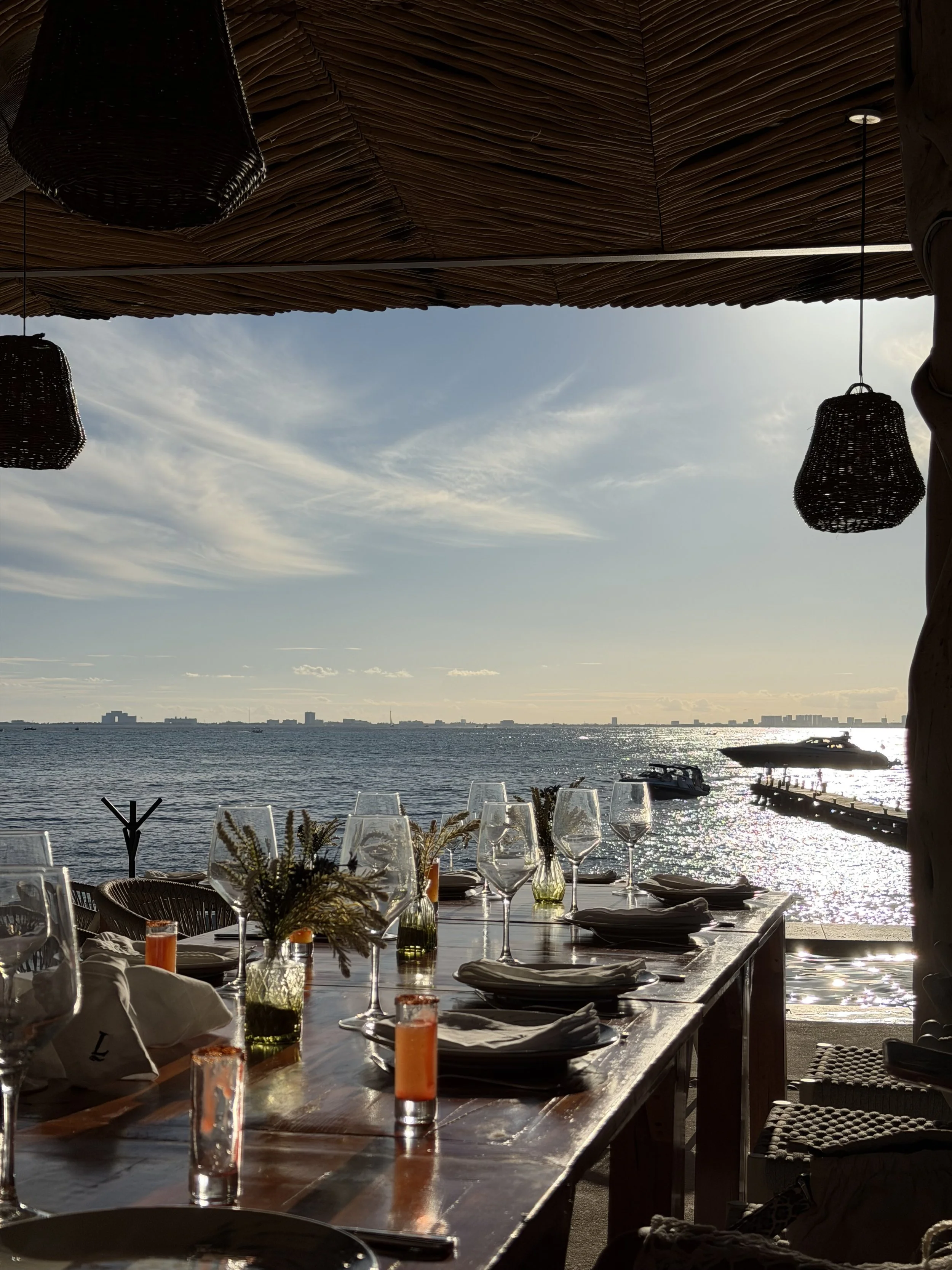 A waterfront dining setup with a long wooden table, wine glasses, plates, and napkins, overlooking a body of water with boats and a distant city skyline, under a thatched roof with hanging wicker lamps.