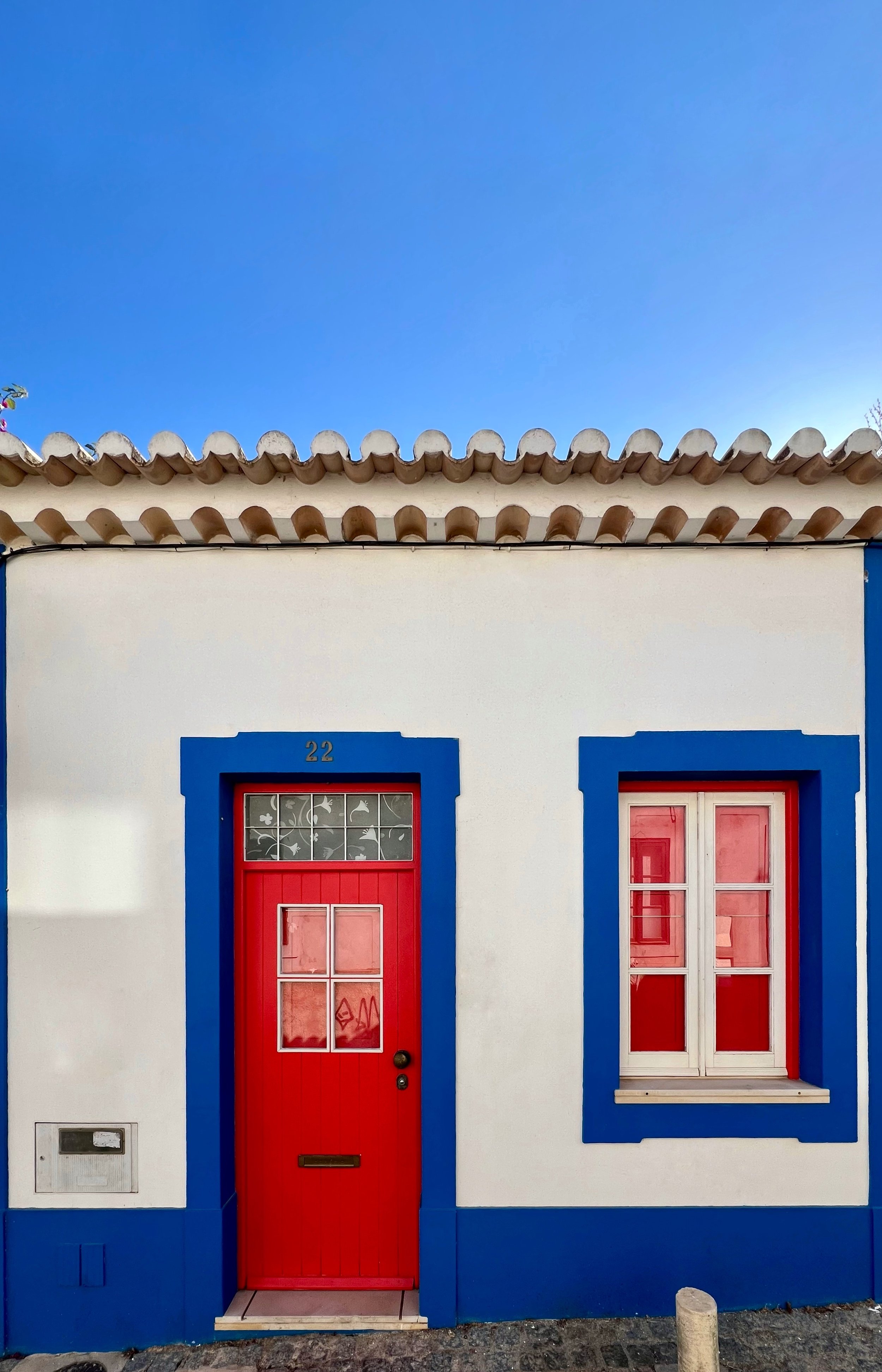 Front view of a house with a white facade, a red door with a window, and a red window framed in blue, under a clear blue sky.