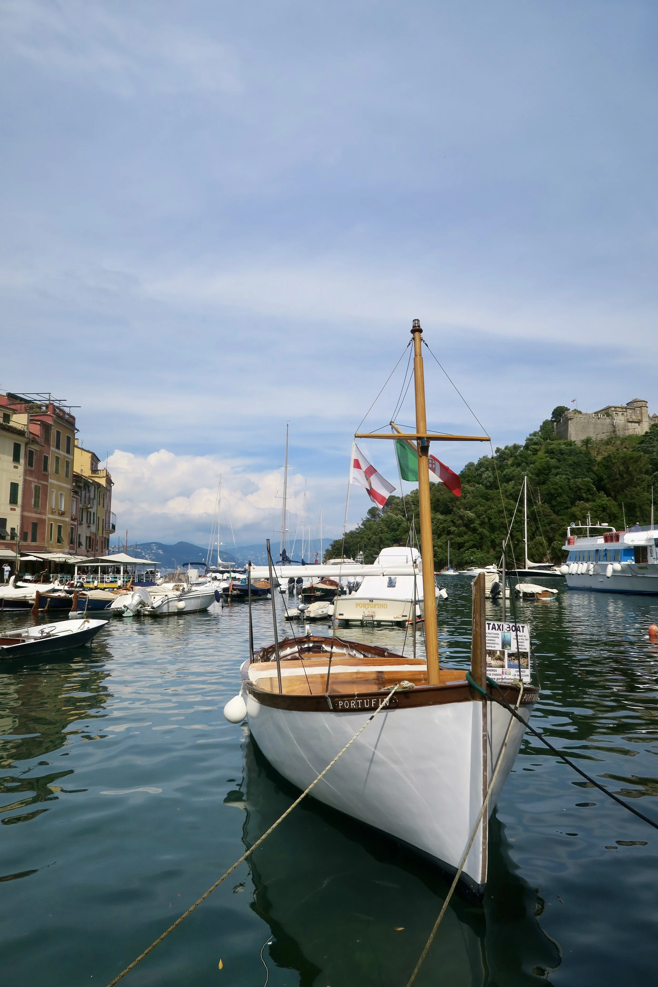 A marina with multiple boats docked in the water, colorful buildings on the left, a hillside with a historic stone fortress in the background, and a clear sky above.