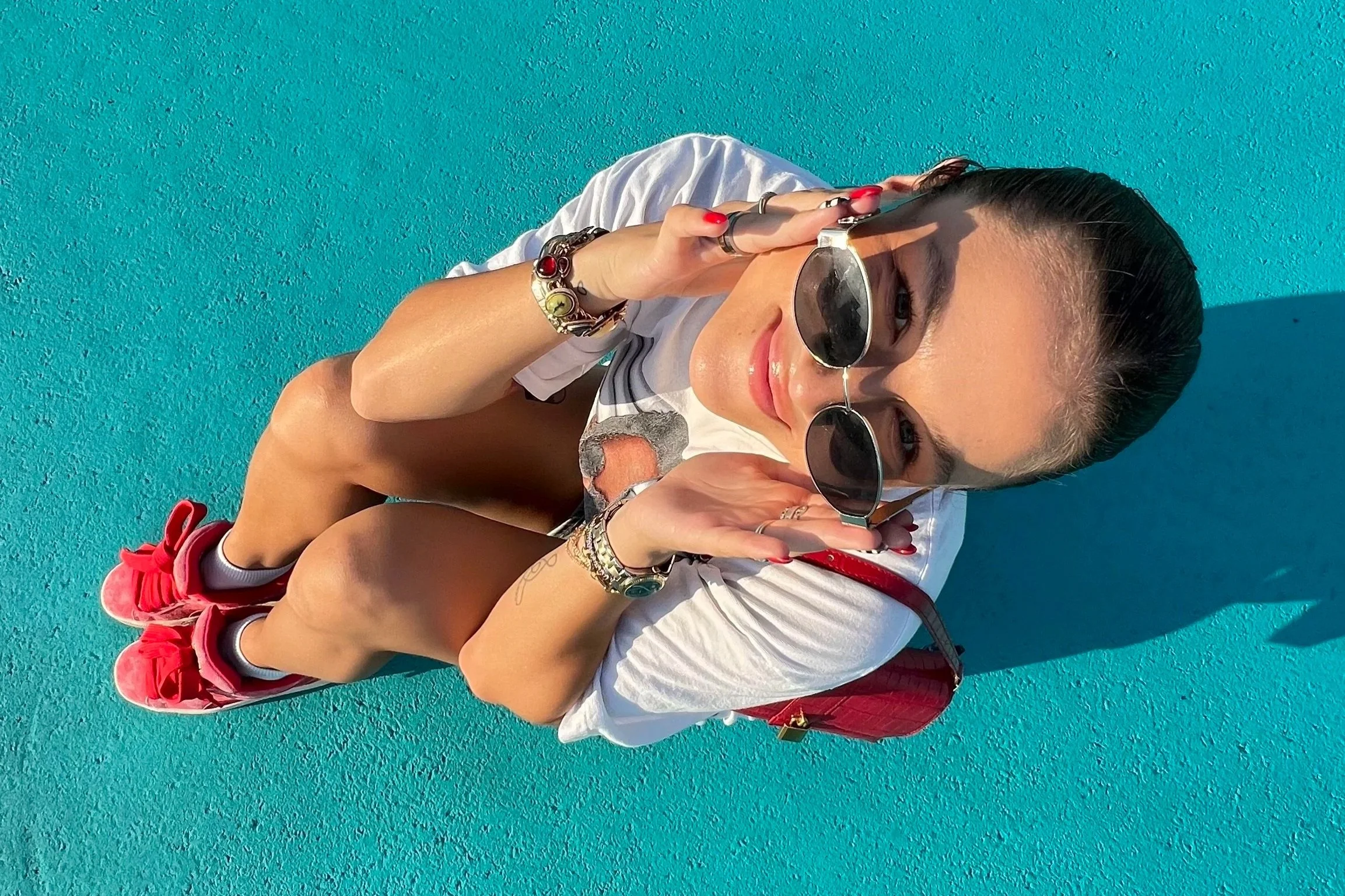 A woman with dark hair wearing sunglasses, a white t-shirt, pink sneakers, and red accessories, sitting on a vibrant blue surface, smiling at the camera.