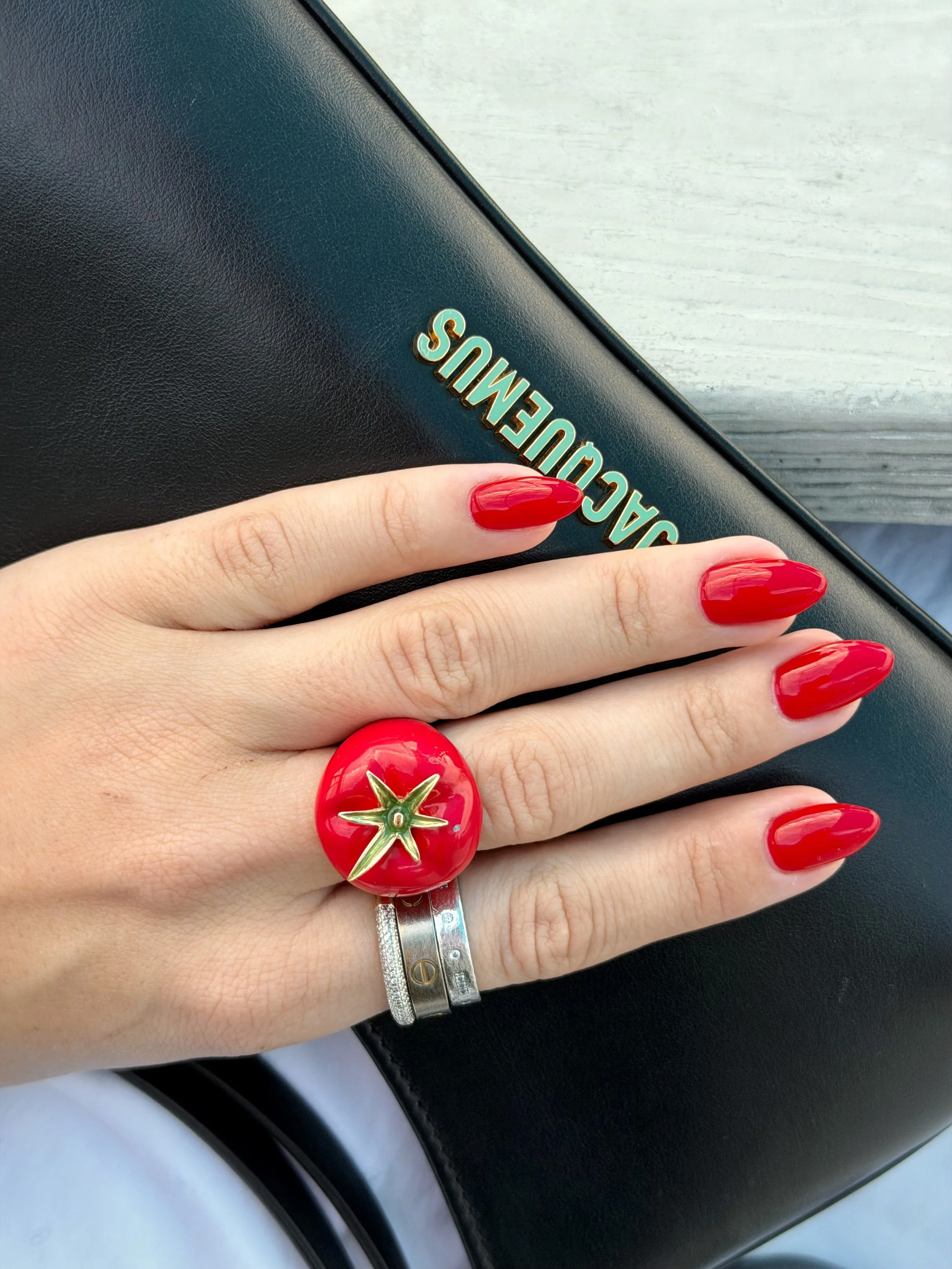 A person's hand with red nail polish and a large tomato-shaped ring resting on a black leather wallet that has a gold and green logo. The person's wrist is adorned with a silver bracelet. The background features a light-colored wooden surface.