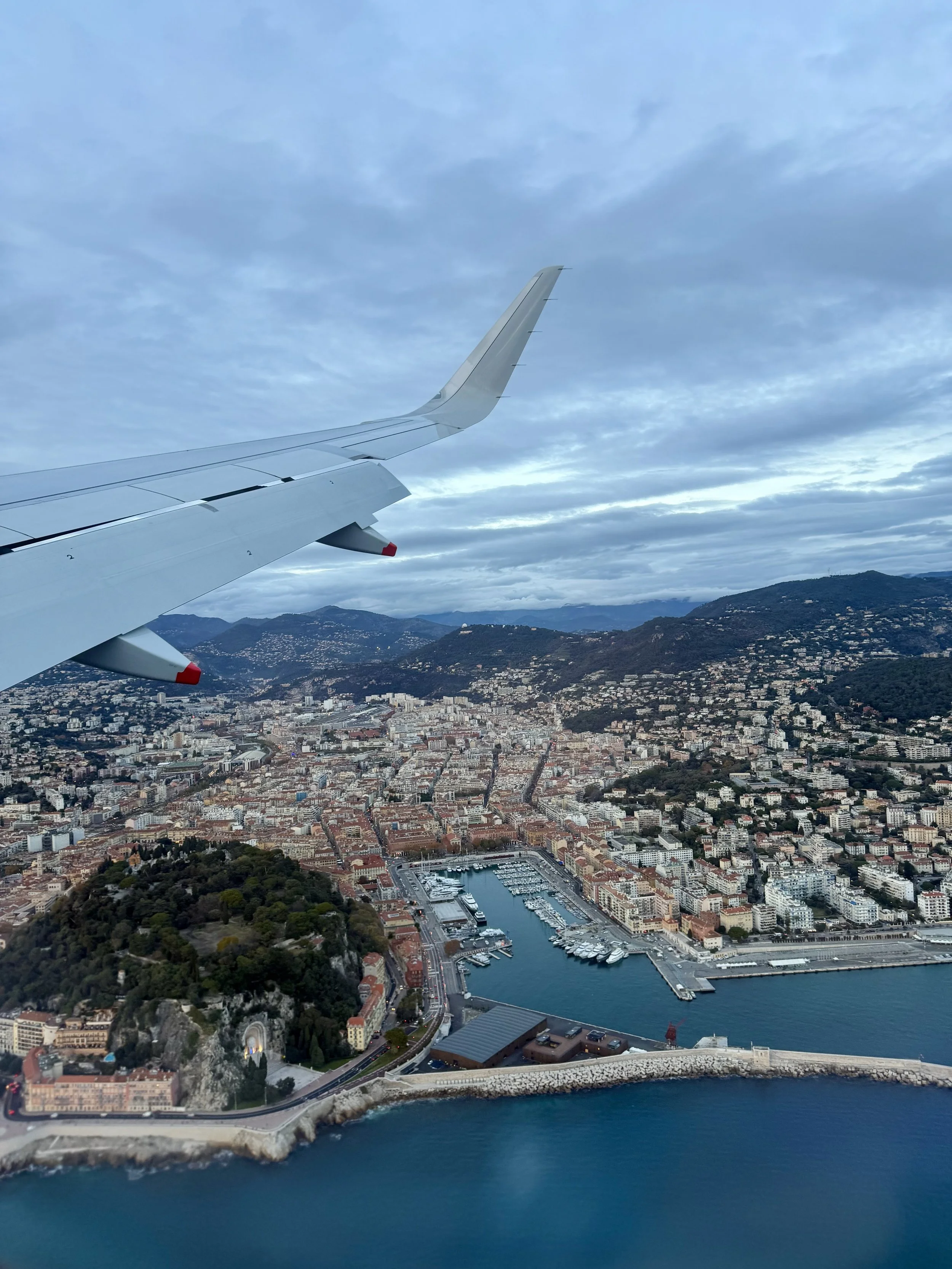 Aerial view of a city with a harbor and coastline, seen from an airplane in flight. The city has dense buildings, roads, and a marina with boats, surrounded by hilly terrain under a cloudy sky.