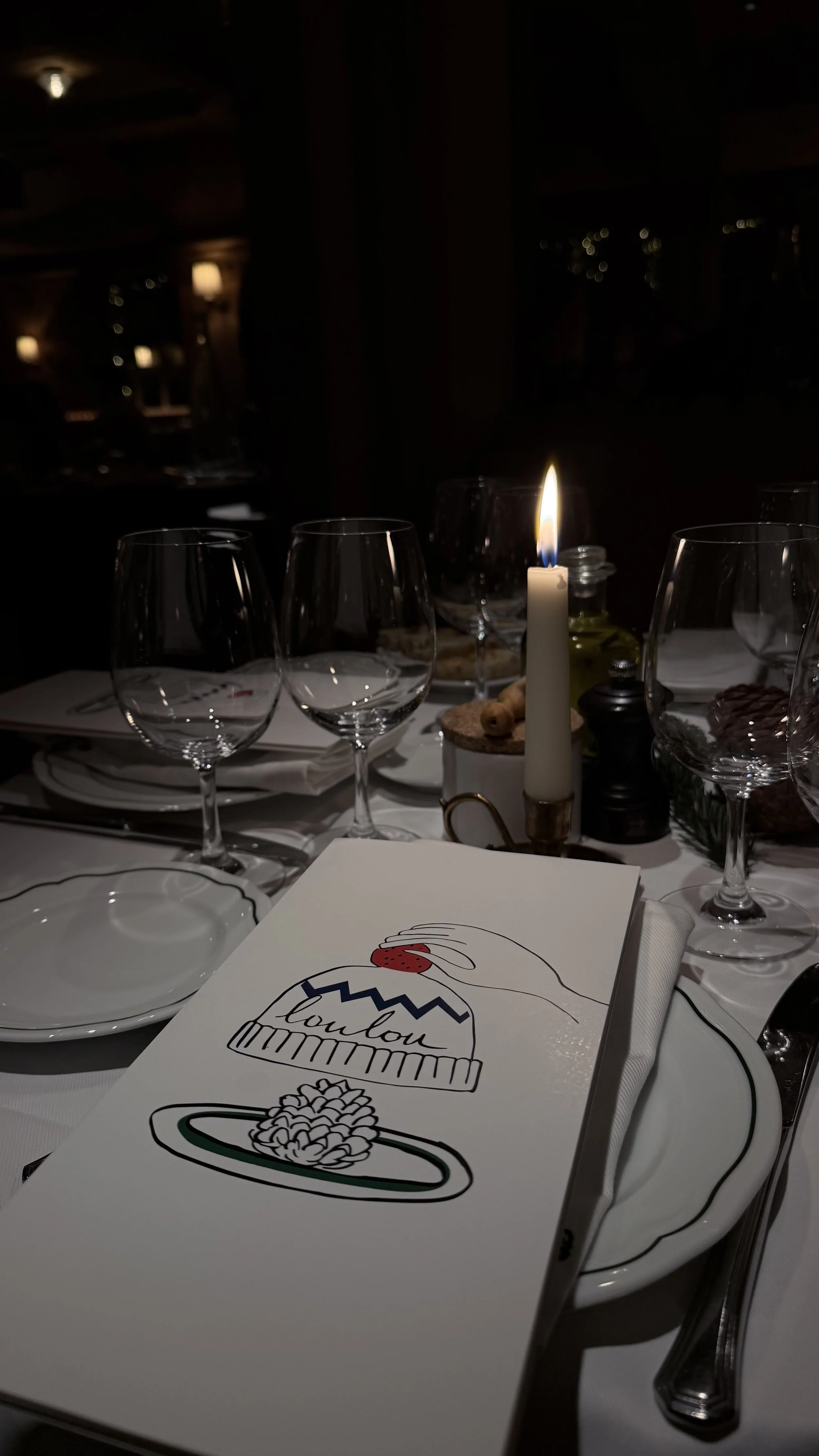 A table set for dinner with wine glasses, a candle, and a menu featuring a drawing of a pasta dish and a chef's hat. The setting appears to be in a dimly lit restaurant.
