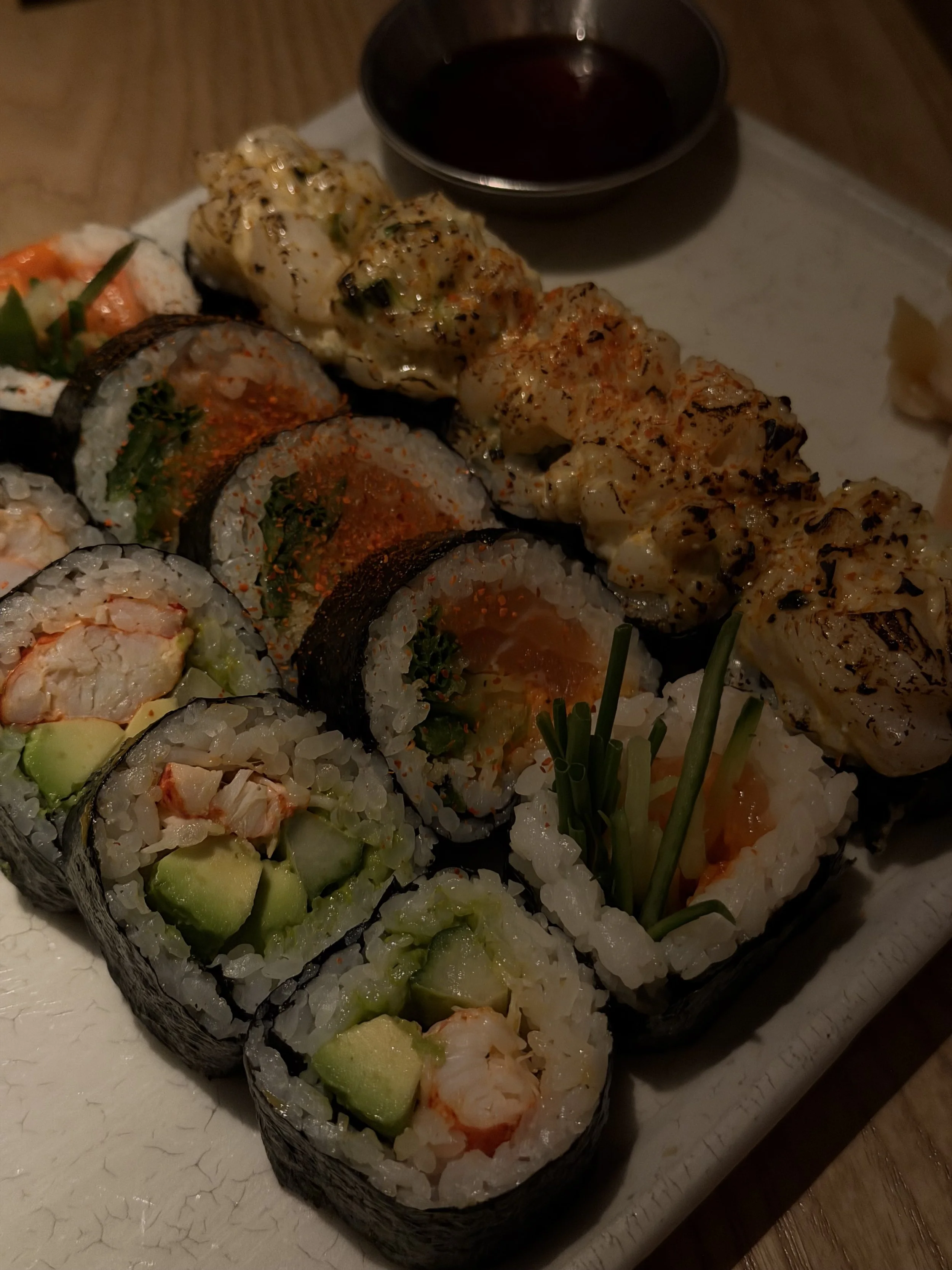 An assortment of sushi rolls with various fillings and toppings on a white serving platter, accompanied by a small dish of soy sauce.