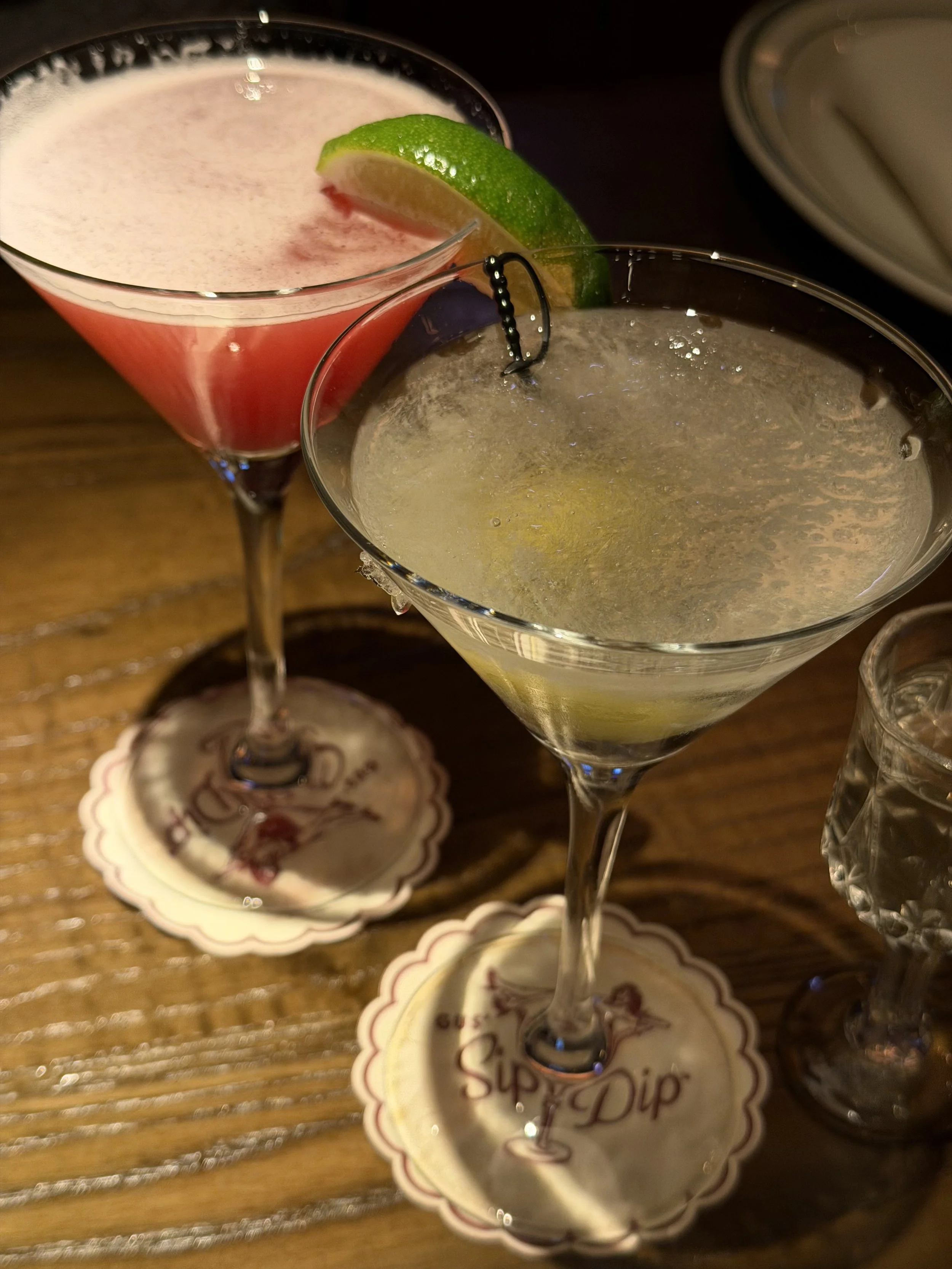 Two cocktail glasses with lime wedges on the rim, placed on a wooden table with decorative coasters.