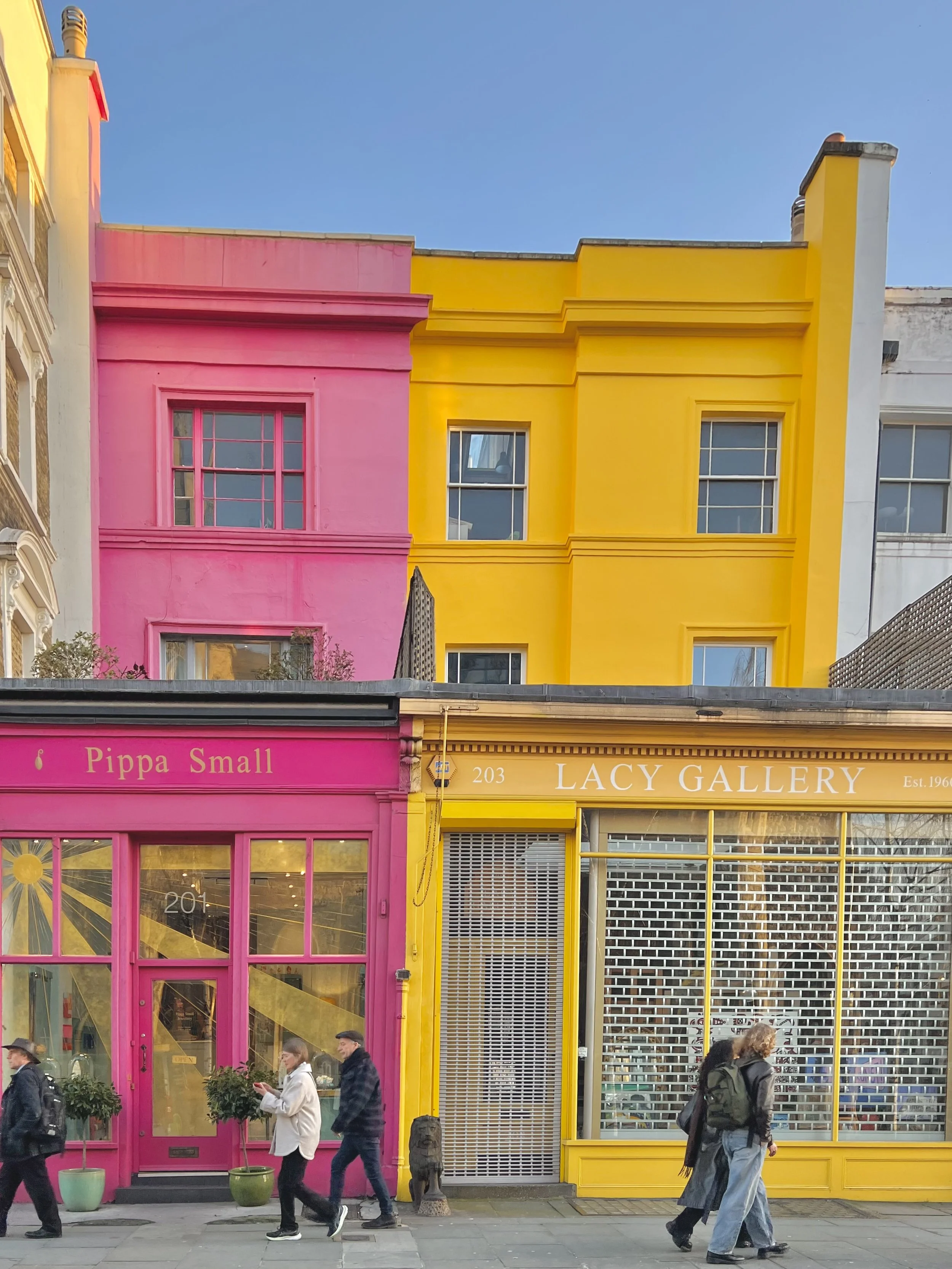 Colorful storefronts with pink and yellow facades on a city street, people walking by, some looking at their phones, trees in front of the stores, and a clear blue sky above.
