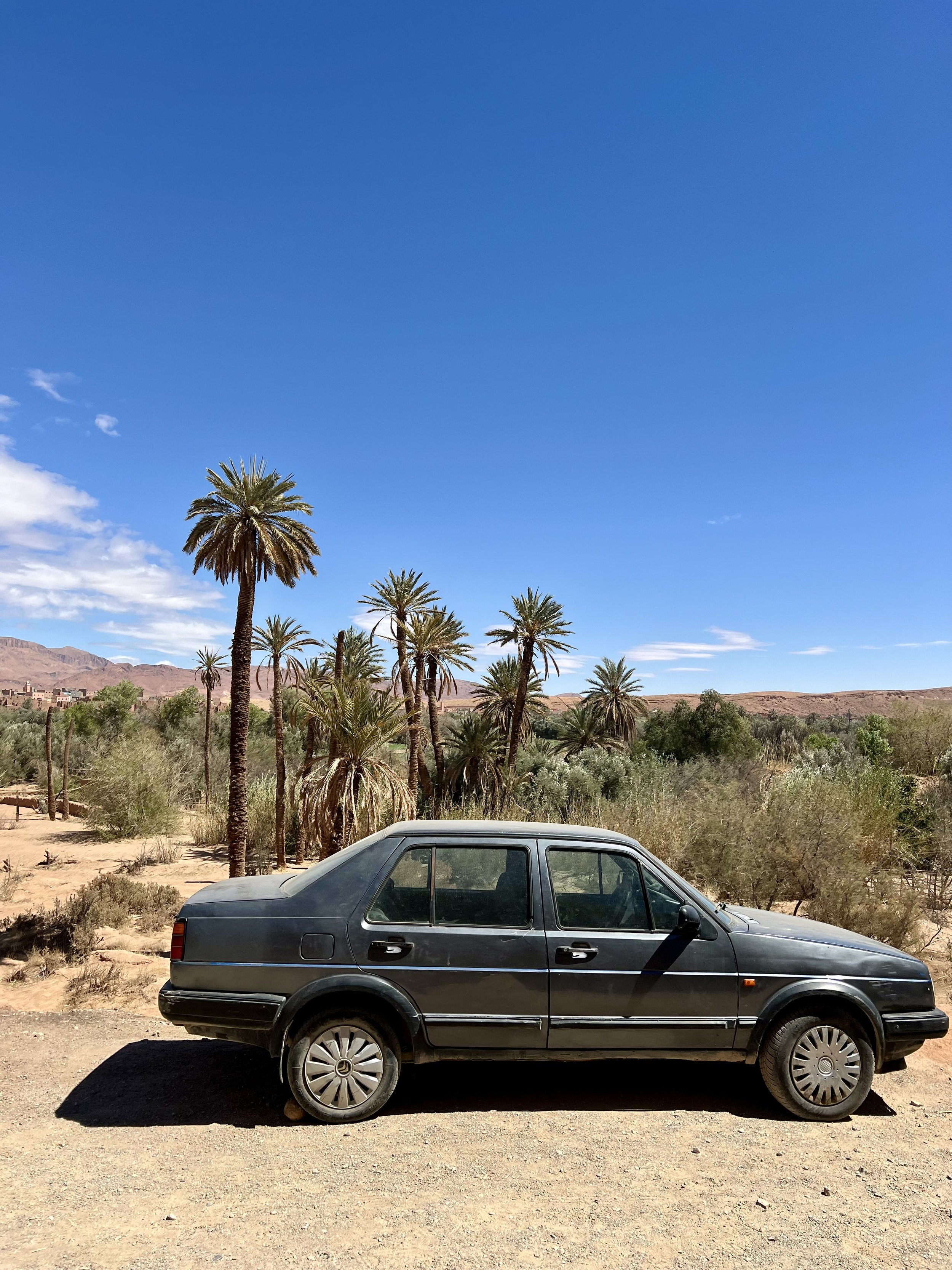 A black classic sedan parked on a dirt road in a desert landscape with palm trees and mountains in the background, under a clear blue sky.