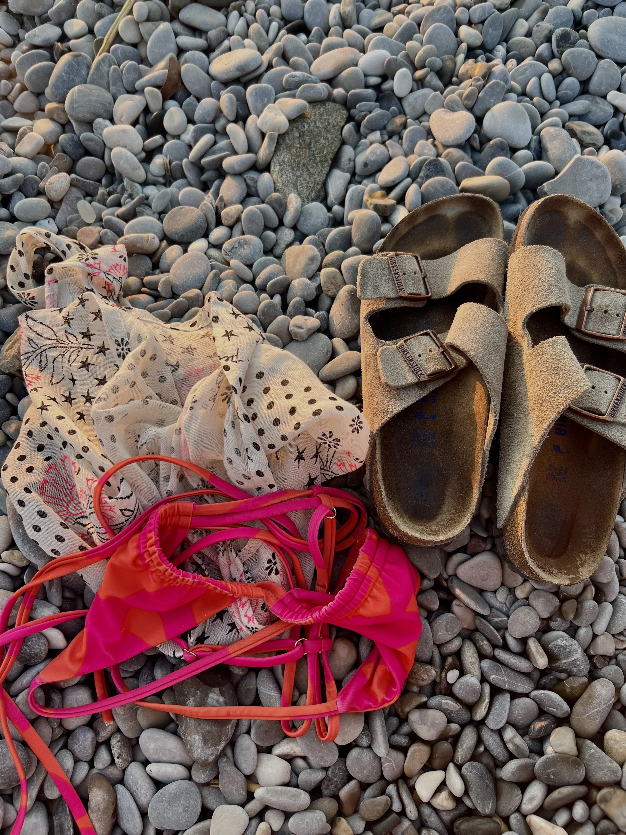A pair of worn sandals, a pink swimsuit, a patterned scarf, and pink safety goggles placed on gray pebbles on a beach.