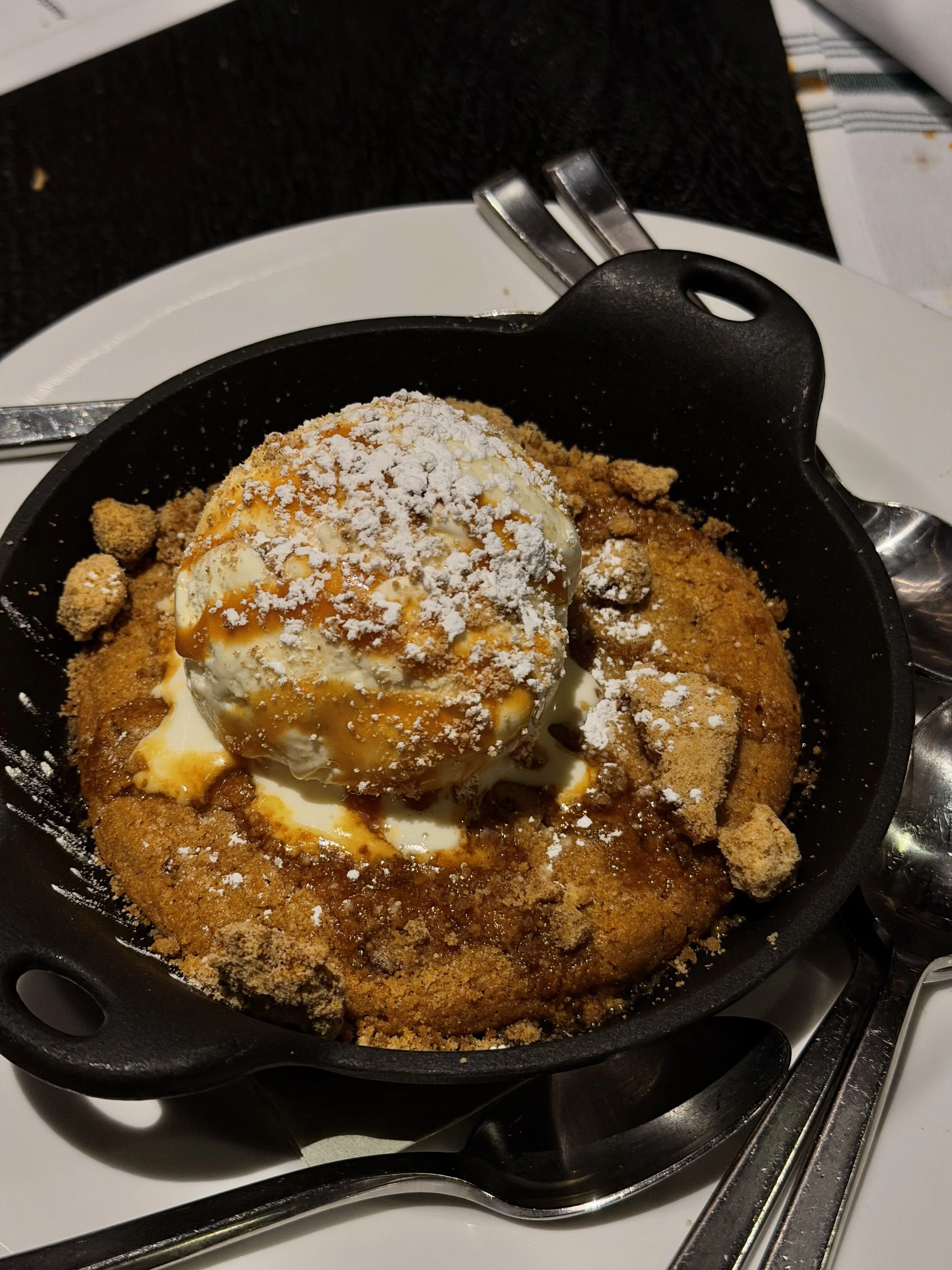 Fried chicken with a scoop of vanilla ice cream topped with caramel sauce and powdered sugar in a black skillet placed on a white plate.