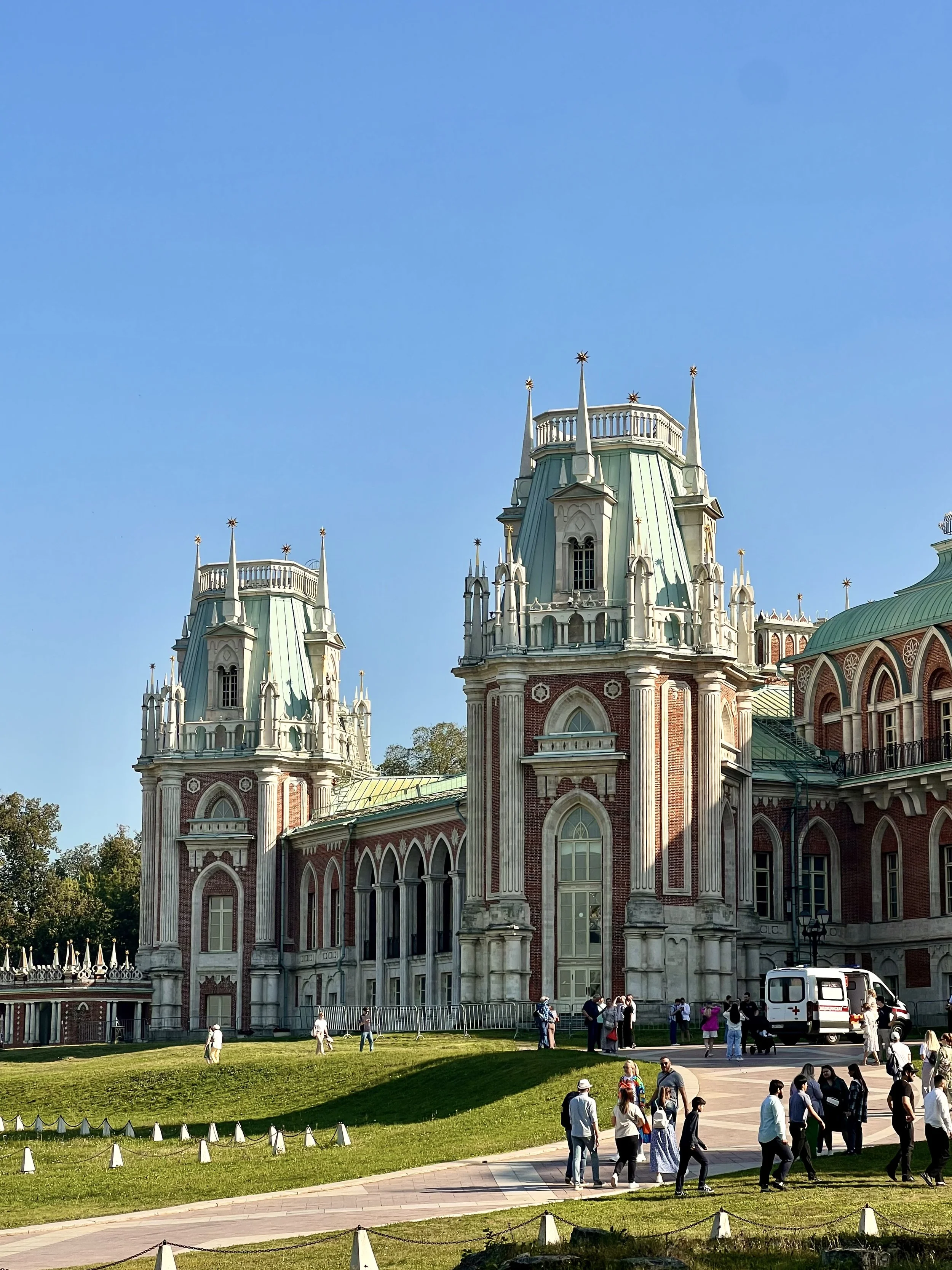 A historic building with spires and ornate architecture, situated in a park area with people walking and gathered on the lawn and sidewalk, under a clear blue sky.
