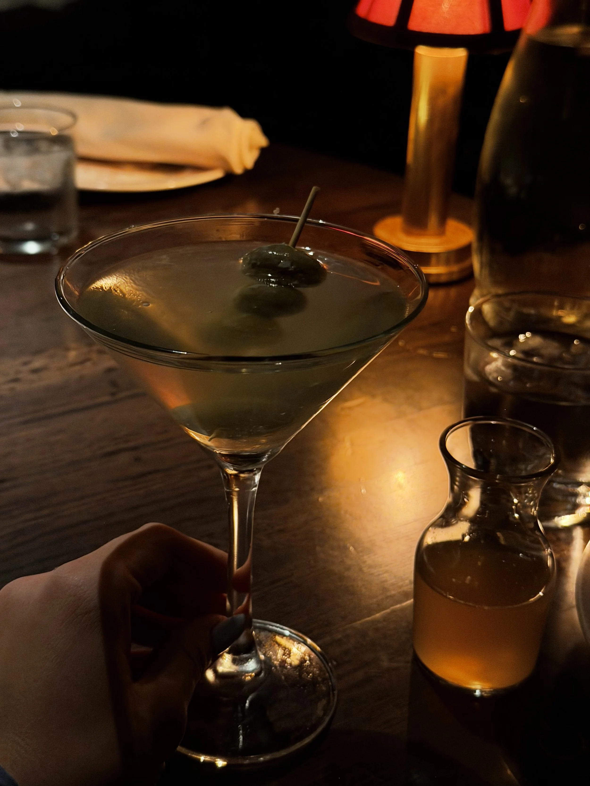 A hand holding a martini glass with an olive garnish, surrounded by a candle, a water glass, and a pitcher on a wooden table in a dimly lit setting.
