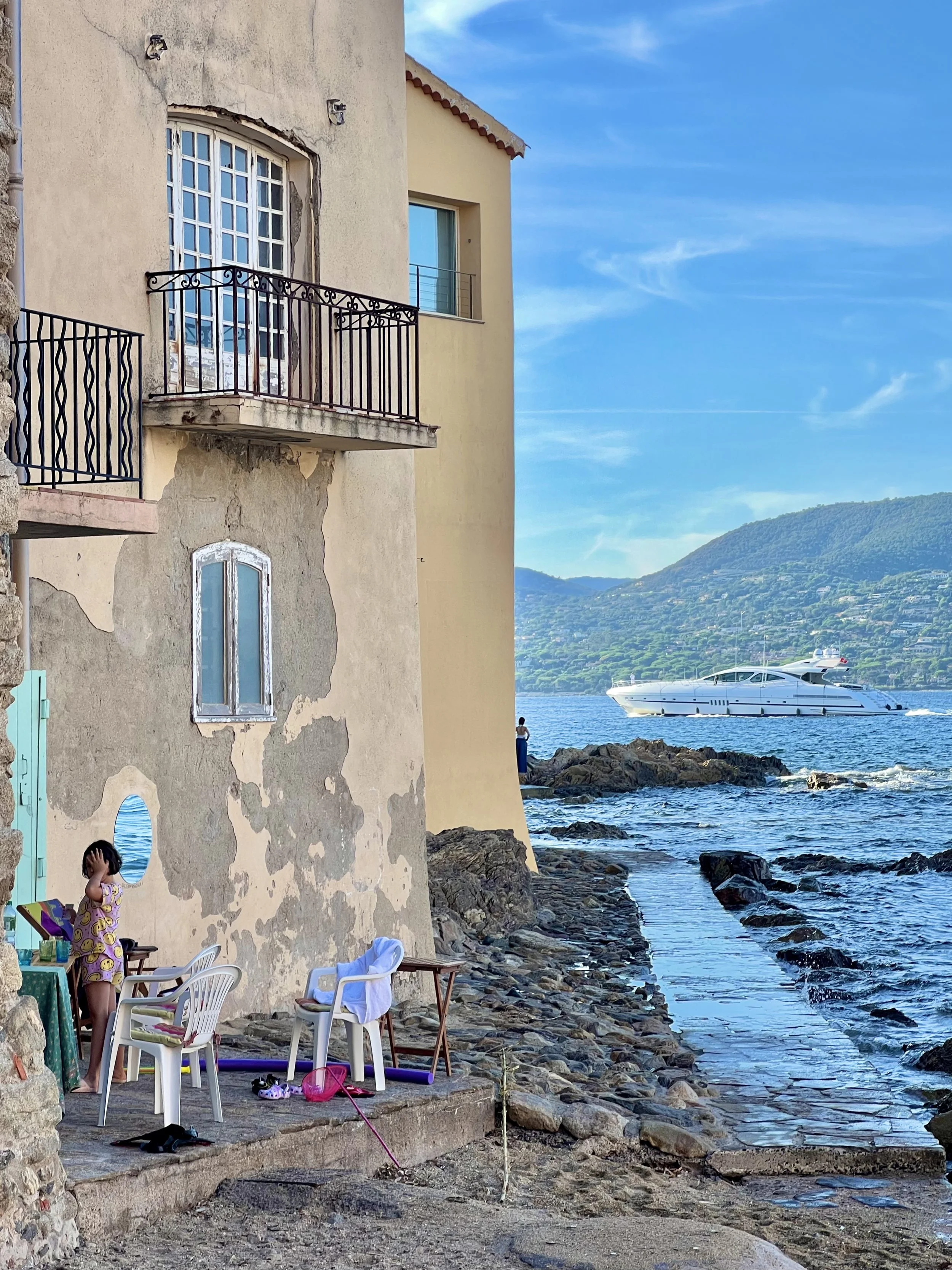 A young girl in pajamas standing near a weathered beige building with an iron balcony, near a rocky shoreline. There are white plastic chairs and a table with a green tablecloth, along with various belongings. In the background, a large white yacht i