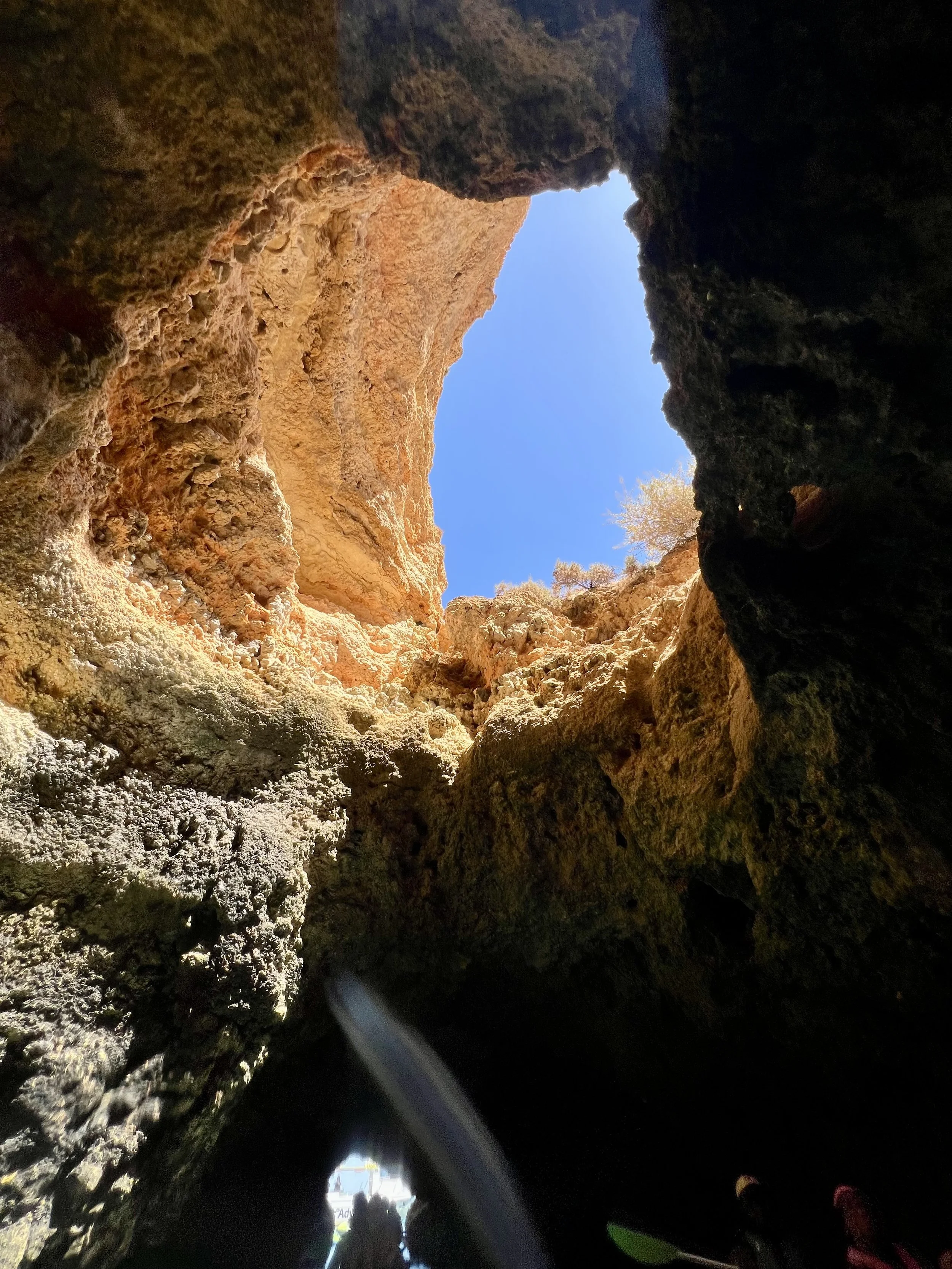 View of the sky through a rocky cave opening, with sunlight illuminating the rough texture of the cave walls and a small tree visible outside the opening.
