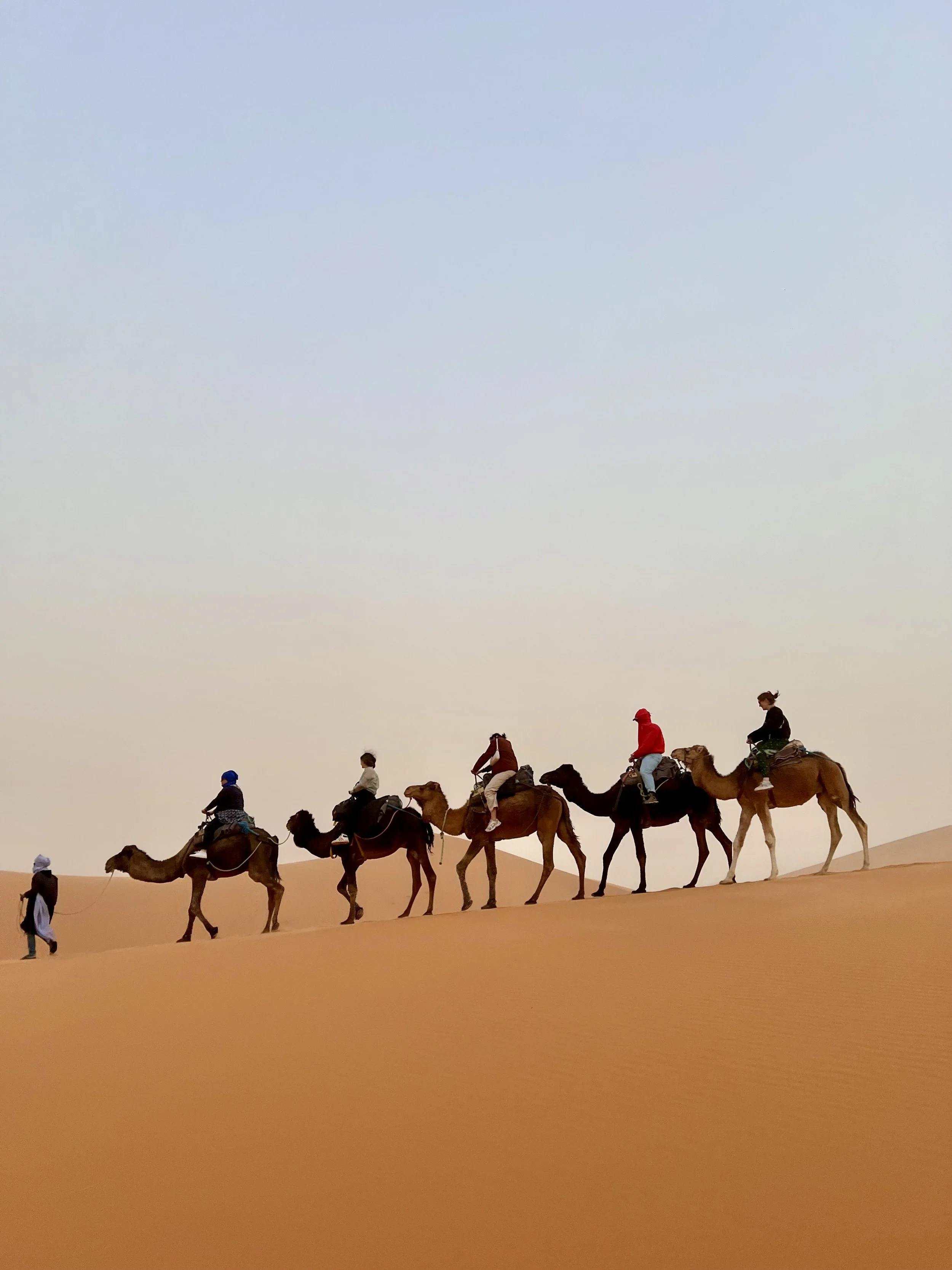 A group of people riding camels across sand dunes in a desert, with some standing and some seated, under a clear sky.