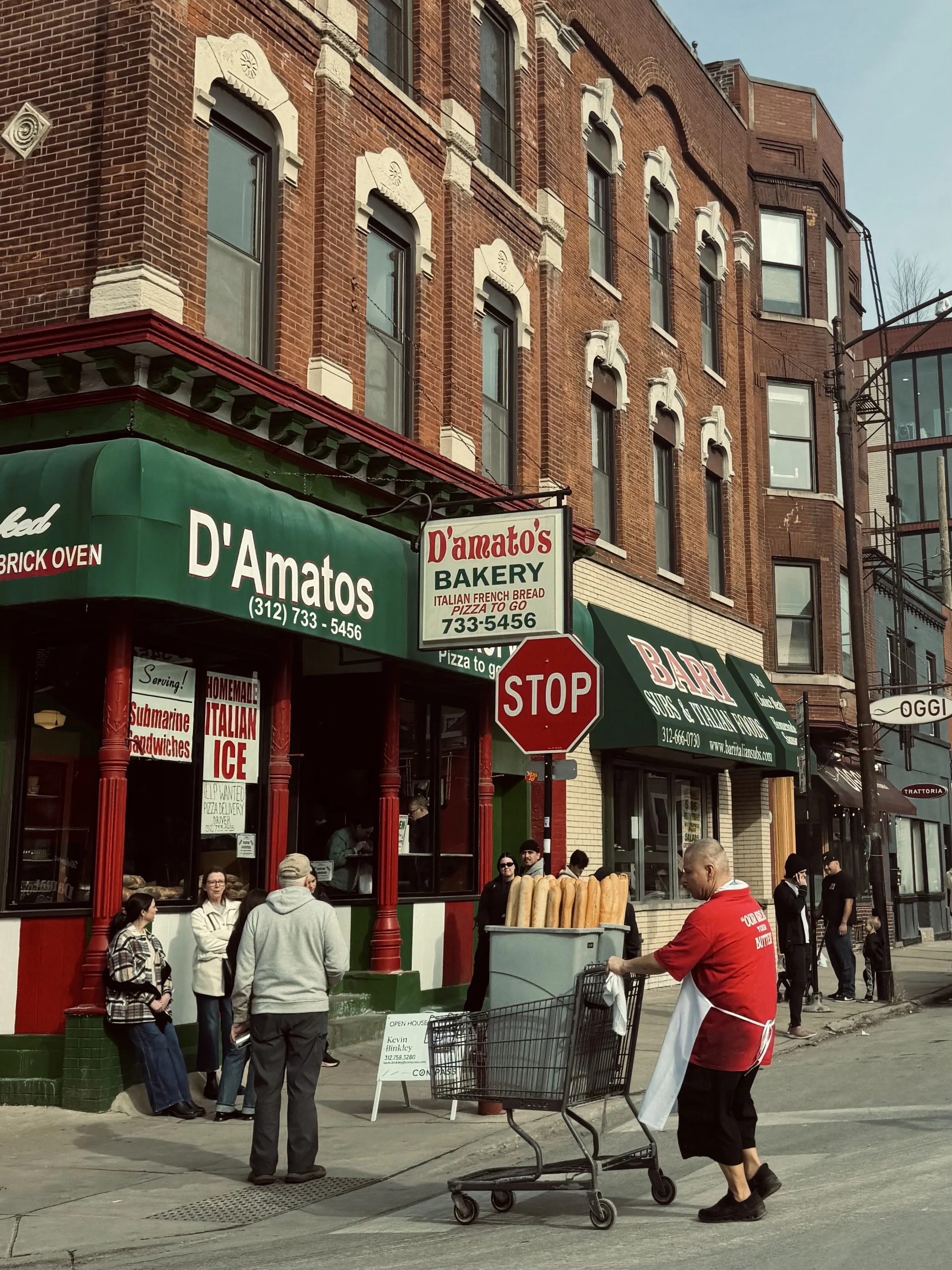 People are standing outside a bakery named D'amato's Bakery, with a green awning and a sign. The bakery offers Italian, French bread, pizza, and sandwiches. Some customers are waiting or shopping, and a worker is pushing a cart full of baguettes outs