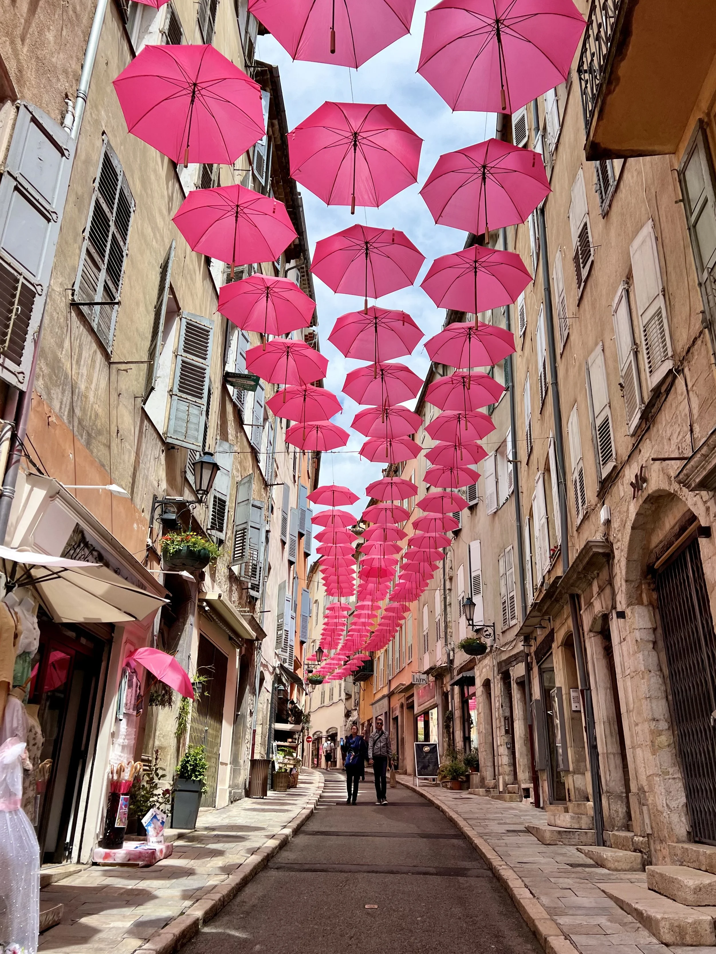 A narrow street decorated with numerous pink umbrellas hanging overhead. The street is lined with pastel-colored old buildings with shuttered windows, and there are a few people walking.