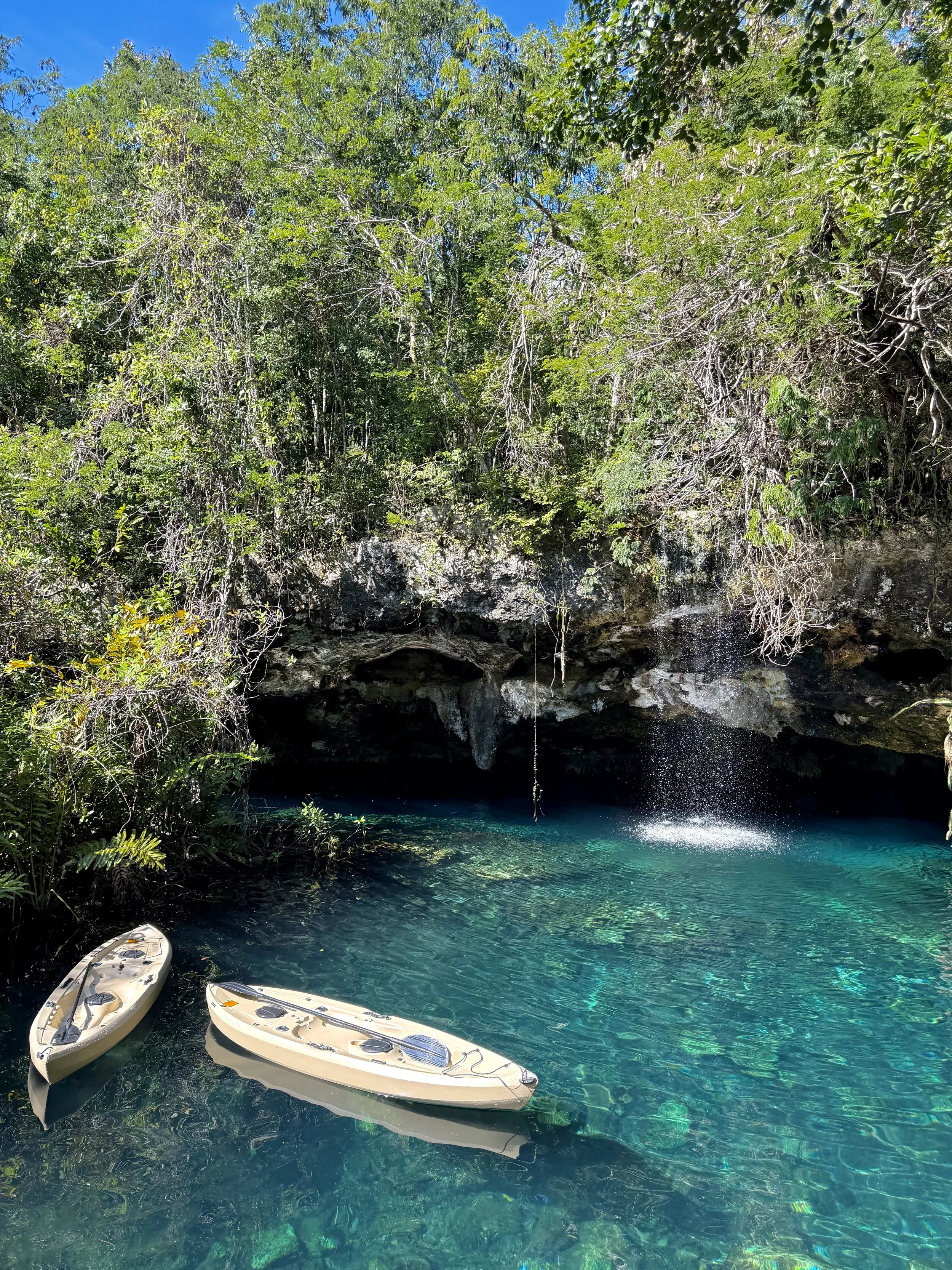 Clear turquoise water with two kayaks, surrounded by lush green trees and rocks, with a small waterfall in the background.
