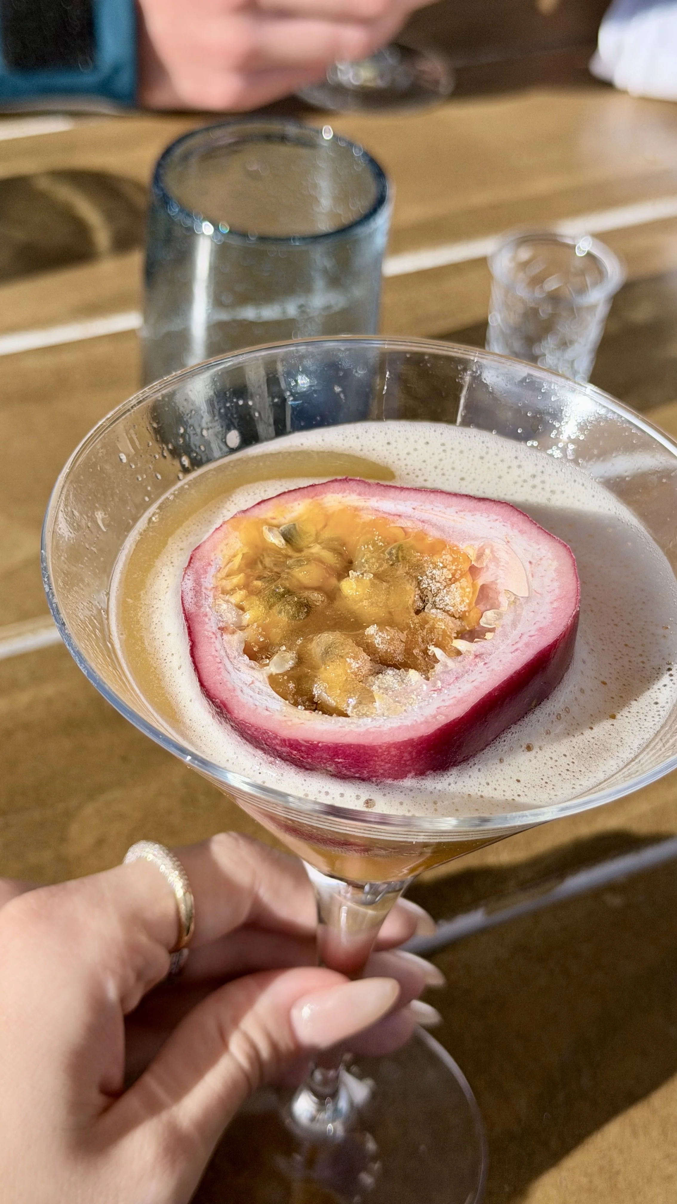 A hand holding a cocktail glass with a pinkish drink topped with a halved passion fruit. The background includes a wooden table with glasses and a person partially visible.
