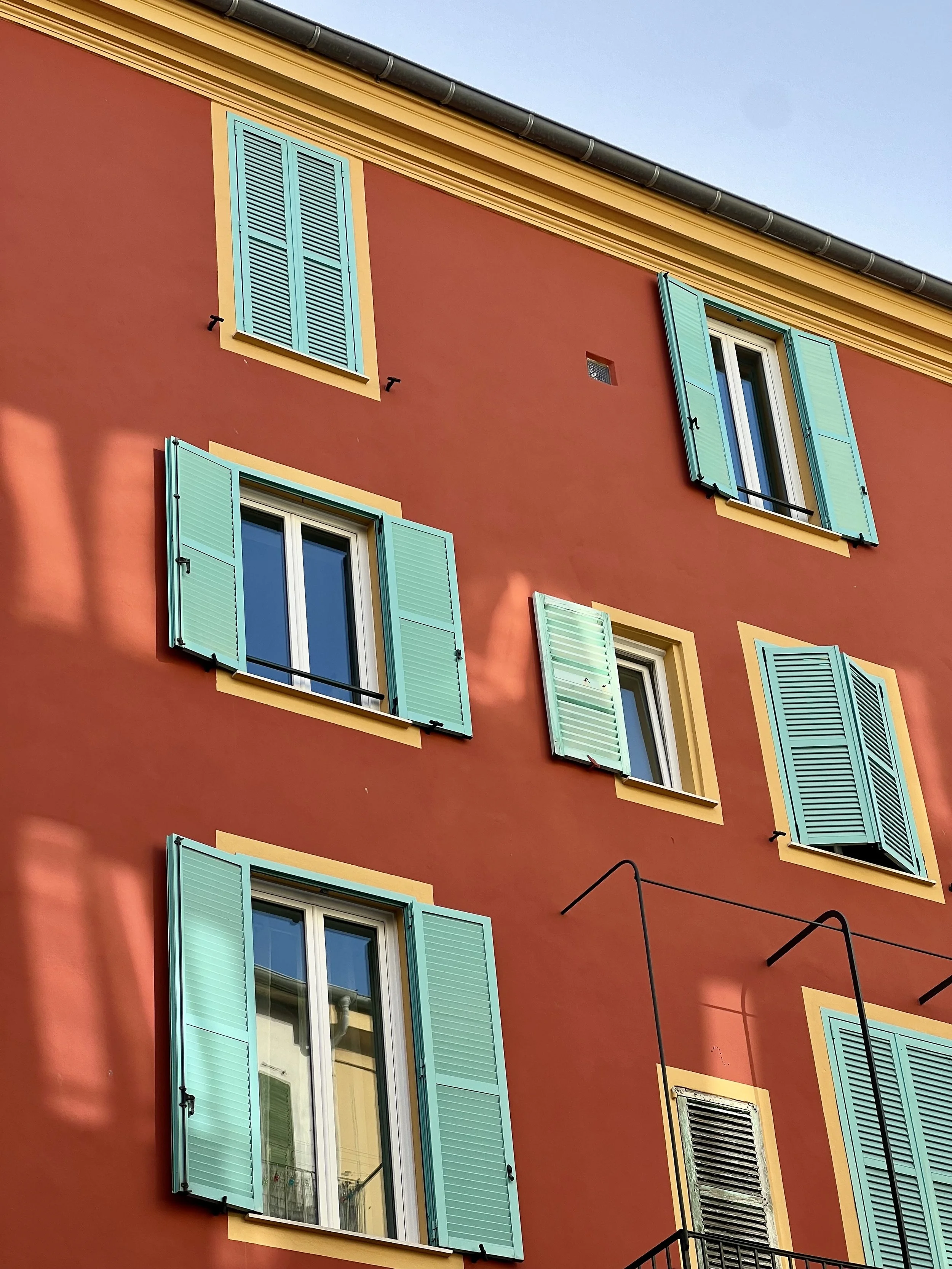 A colorful building with red walls, yellow trim, and turquoise window shutters. The building has multiple windows, some open and some closed, with black metal bars. The sky is clear and blue.