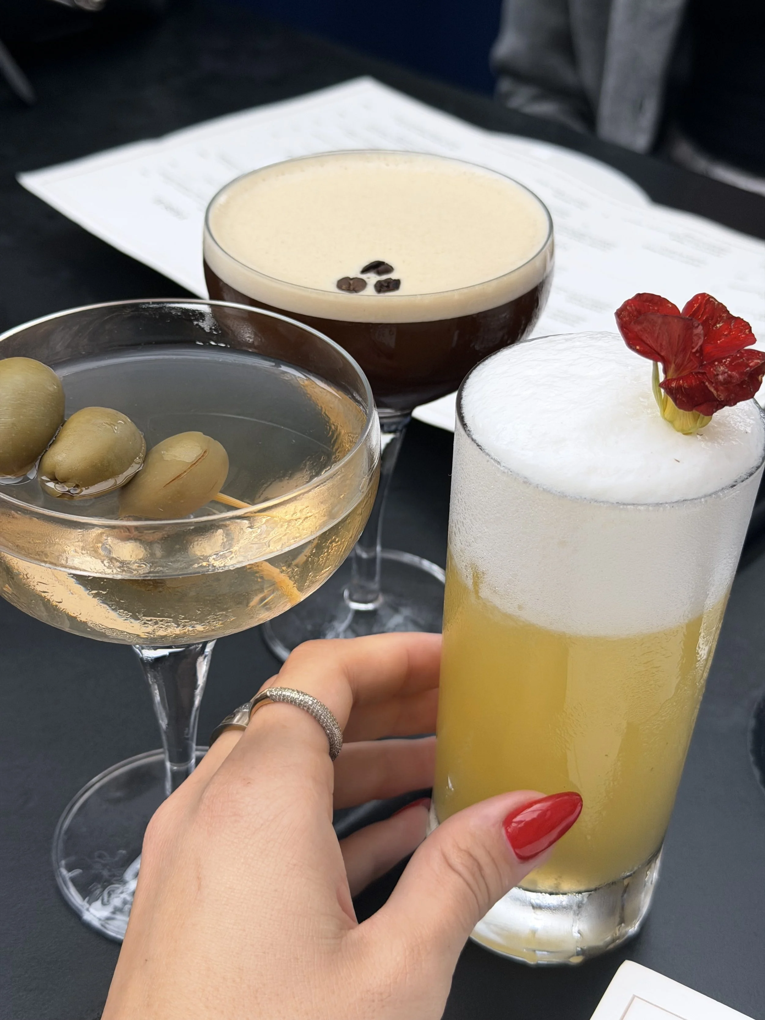 Person holding a tall glass of yellow cocktail with a red flower garnish, three other drinks in glasses behind, one with coffee beans and another with olives, on a black table.