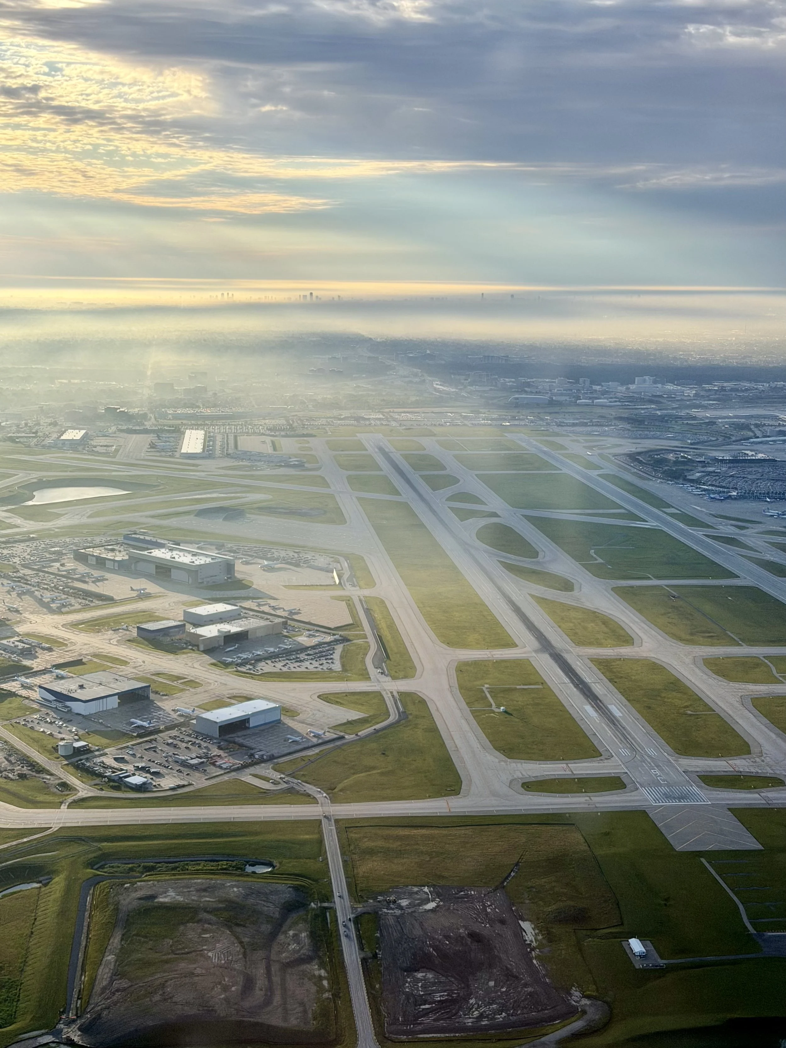 An aerial view of an airport with multiple runways and terminals, surrounded by green fields and some facilities. The sky is cloudy with soft sunlight breaking through.