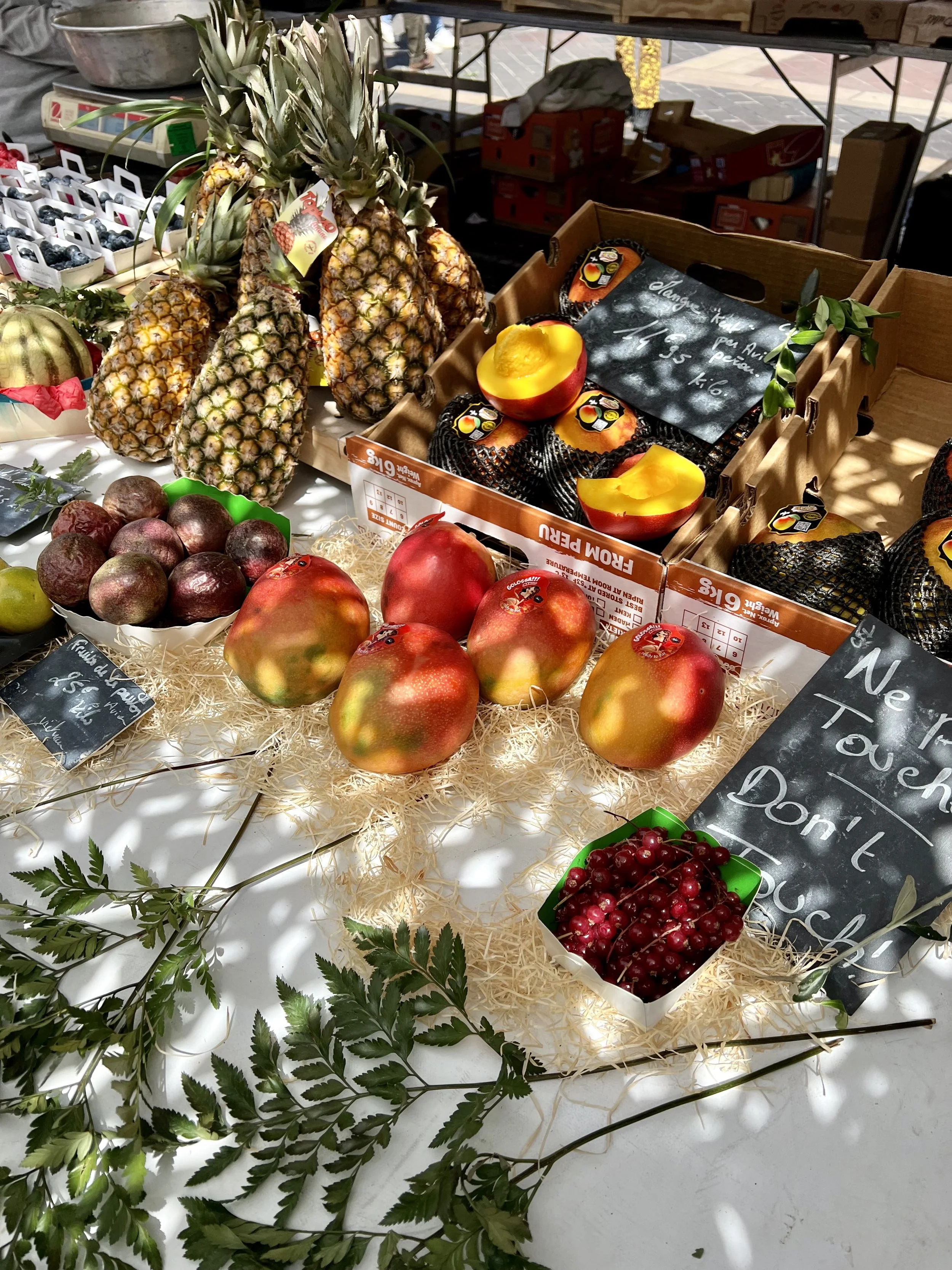 Fresh pineapples, pomegranates, cherries, and peaches displayed on a table at a market, with surrounding green leaves and a handwritten chalkboard sign.