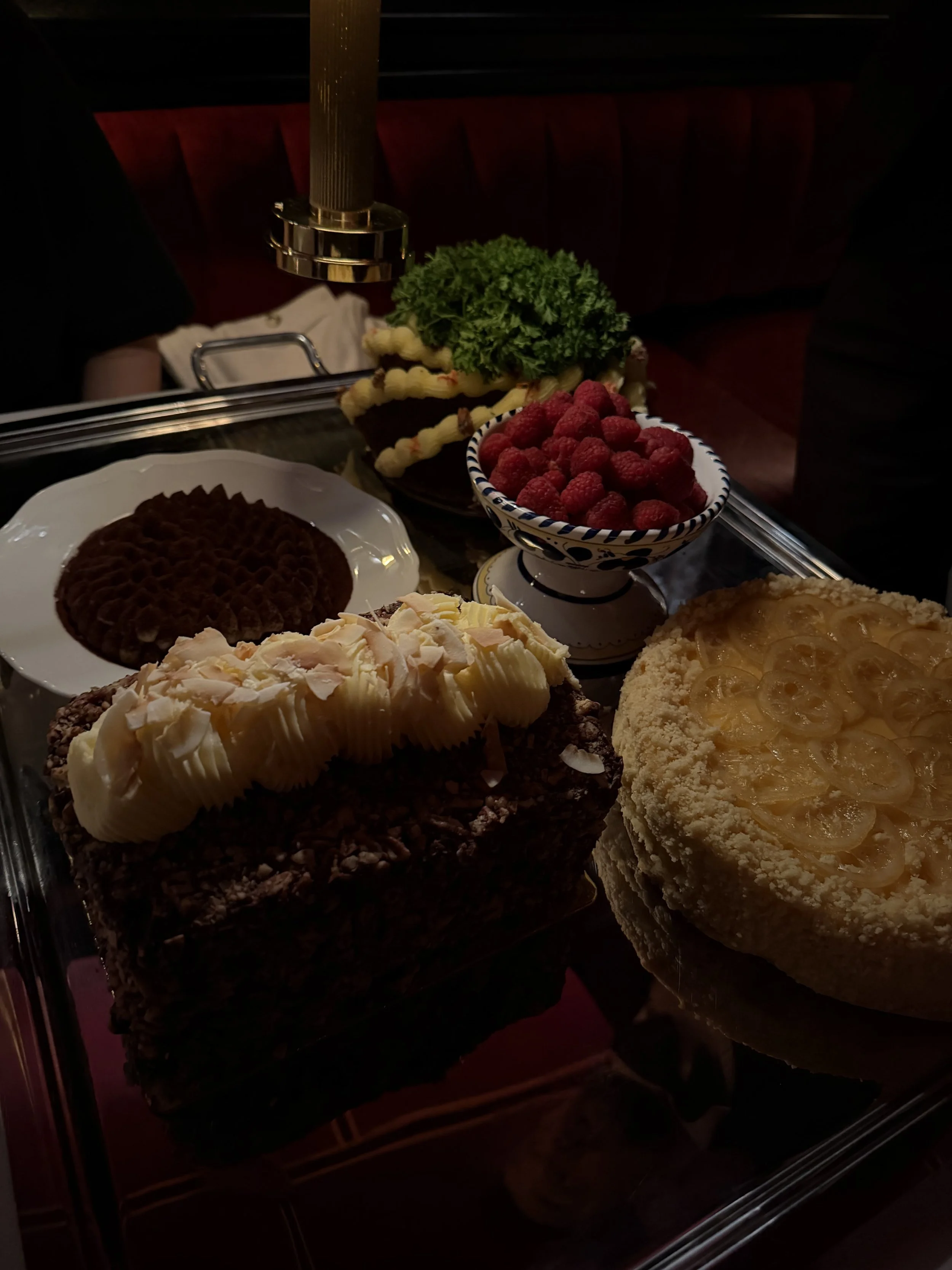 A variety of desserts on display, including a chocolate cake topped with white icing and coconut flakes, a lemon tart with sliced lemon topping, a bowl of fresh raspberries, and a layered cake with yellow frosting, along with a chocolate cake decorat