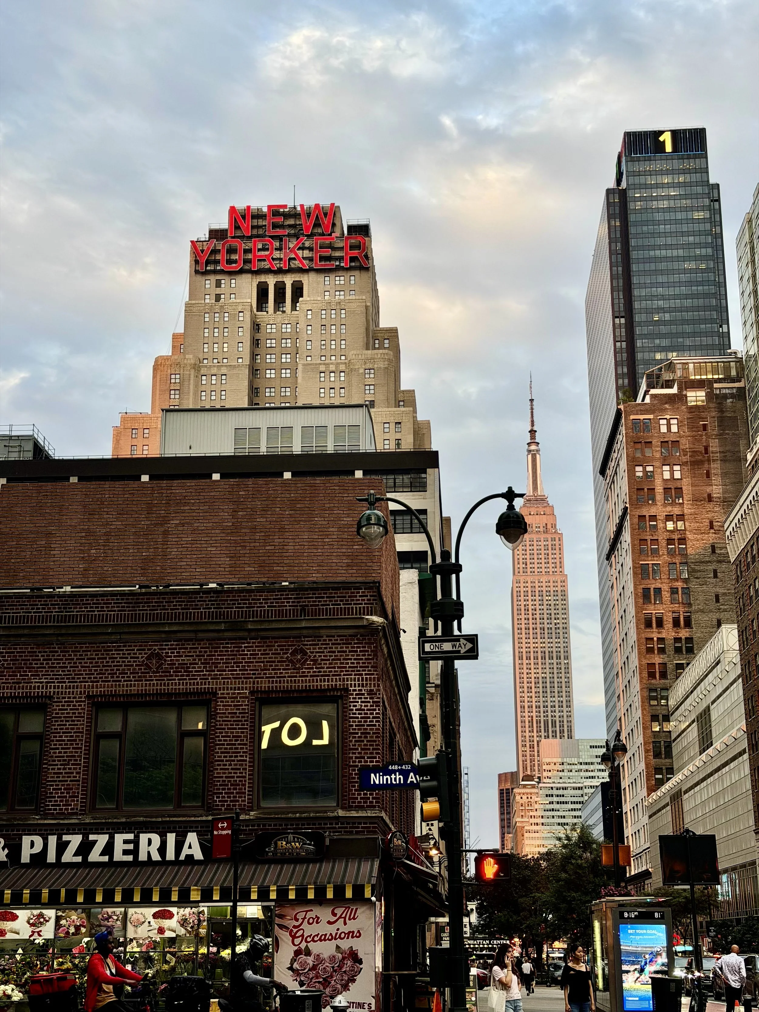 Street scene in New York City with the Empire State Building in the background, a New Yorker sign on a tall building, pedestrians on the sidewalk, storefronts, and traffic lights.