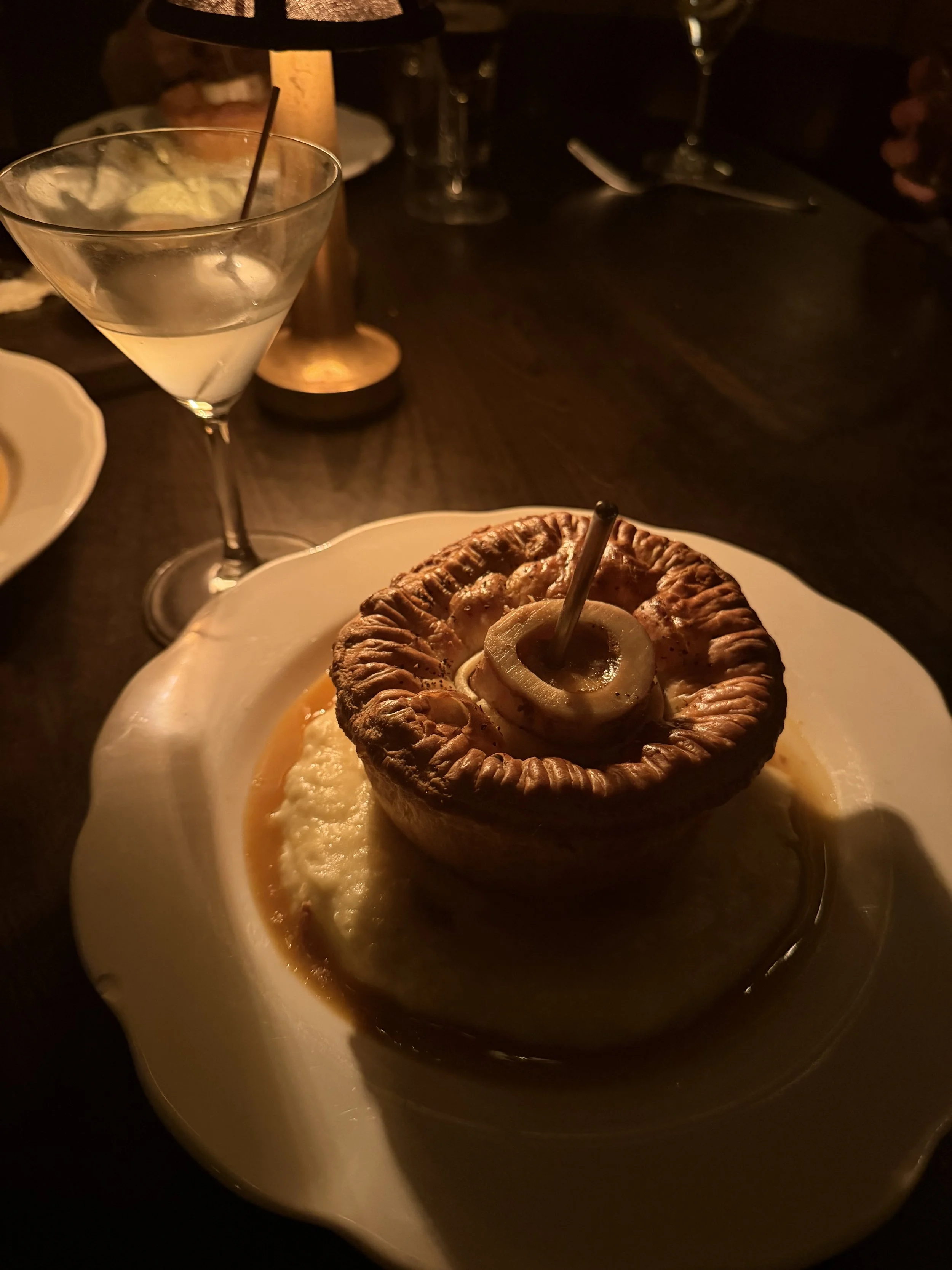A savory baked pastry with a crust, filled with cheese and topped with cooked onion rings, served on a white plate. In the background, a glass of cocktail with a lime wedge and straw is visible in a dimly lit setting.