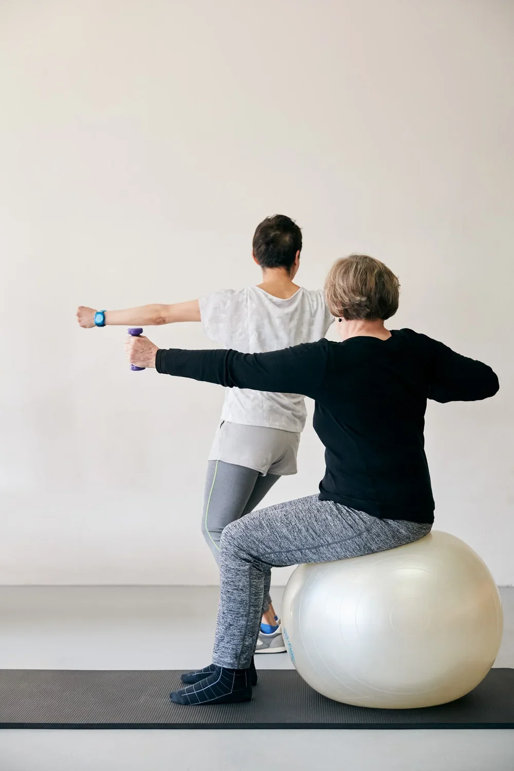 A woman sitting on a large exercise ball and an instructor assisting her with arm stretches in a fitness or physical therapy setting.
