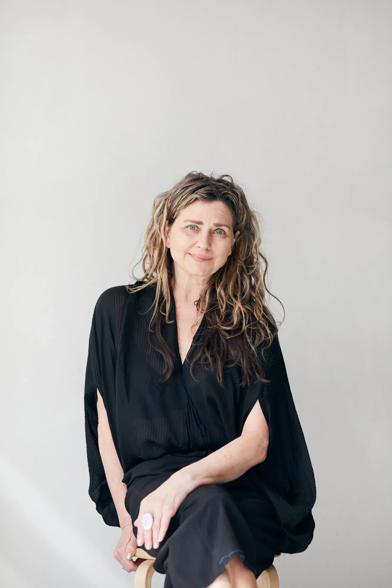 A woman with wavy, light brown hair and blue eyes is sitting on a wooden stool against a plain, light gray background. She is wearing a black, pleated dress and has a subtle smile on her face. Her arms are crossed, with one hand resting on her knee and the other on her arm, displaying a large ring on her finger.