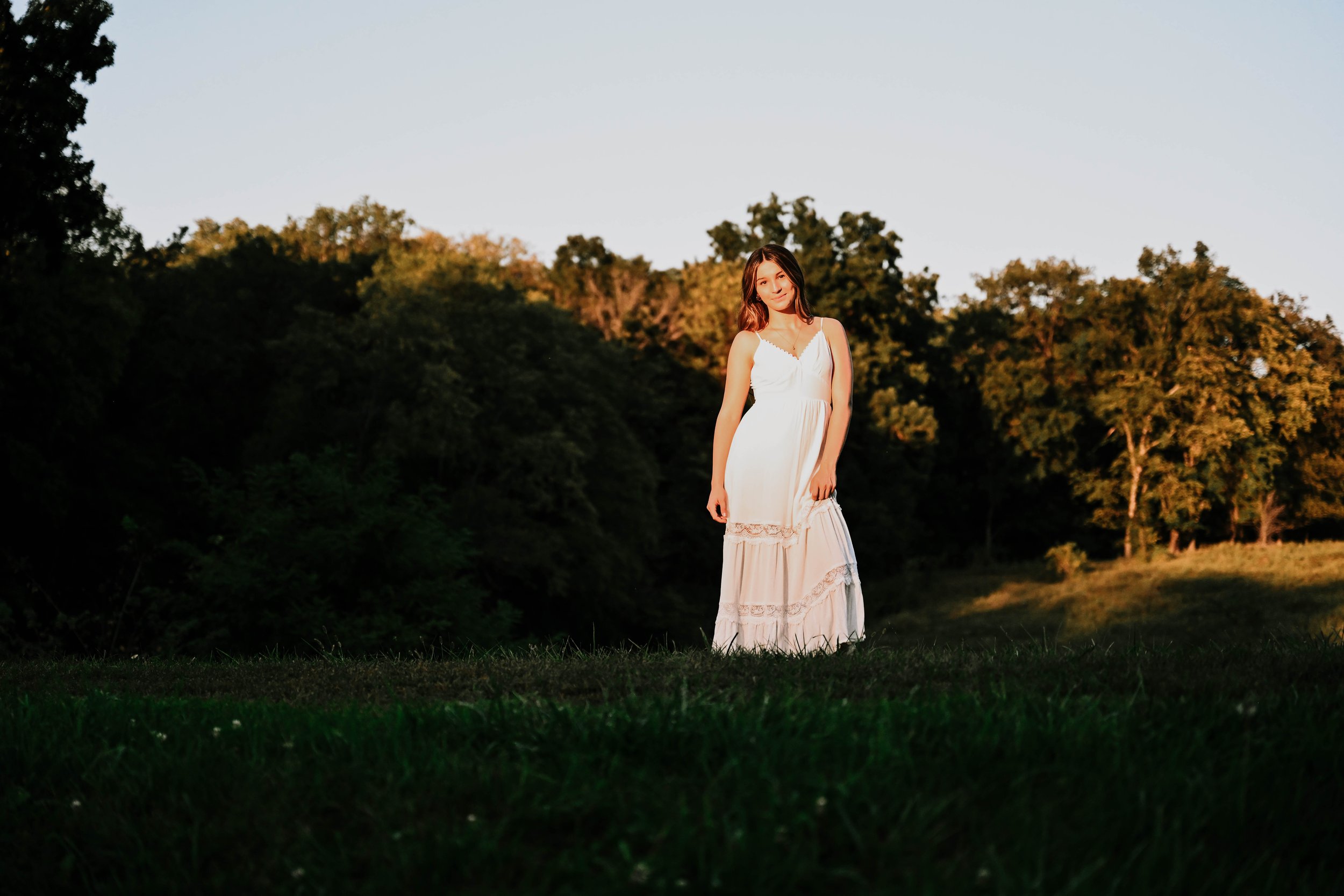 A young woman wearing a white dress standing outdoors during sunset with trees in the background.