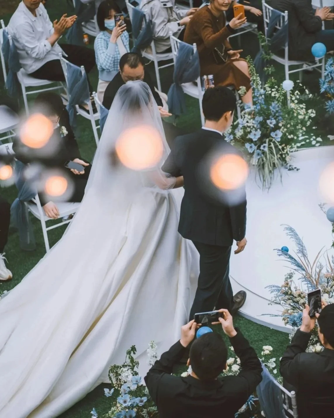 A bride and groom walking down the aisle at their wedding ceremony, surrounded by seated guests taking photos.