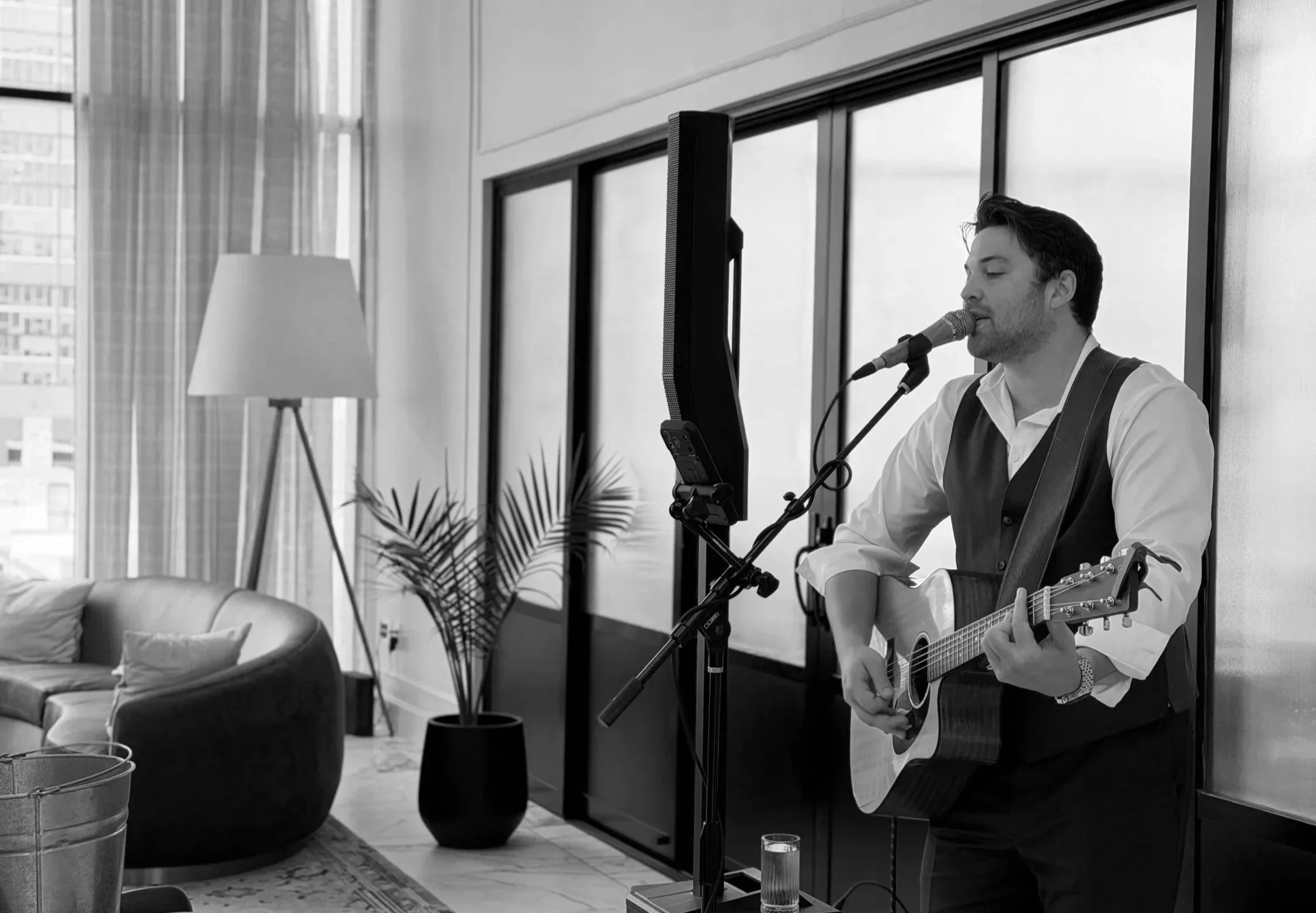 A man dressed in a vest and white shirt playing an acoustic guitar and singing into a microphone in a modern living room with large windows, a sofa, a tall floor lamp, and a potted plant.