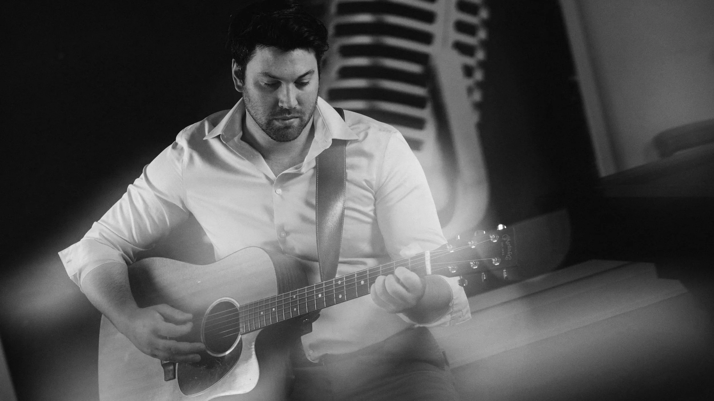 A young man with dark hair and facial hair, wearing a light-colored button-up shirt, plays an acoustic guitar. He is seated indoors, with a background that includes a vent or louvered panel.