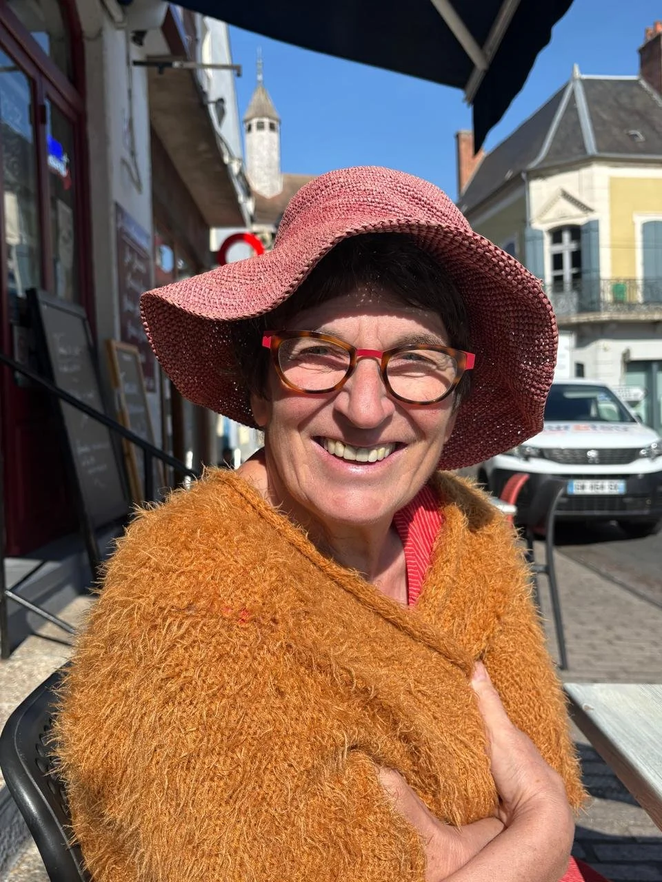 Een lachende vrouw draagt een roze strooien hoed, rode brillen en een oranje, zachte jas, zit buiten op een terras op een zonnige dag.