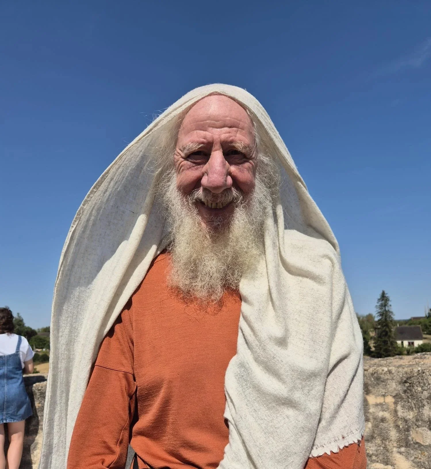 Een oudere man met een witte baard en witte hoofddoek die glimlacht, buiten op een dag met een blauwe hemel.