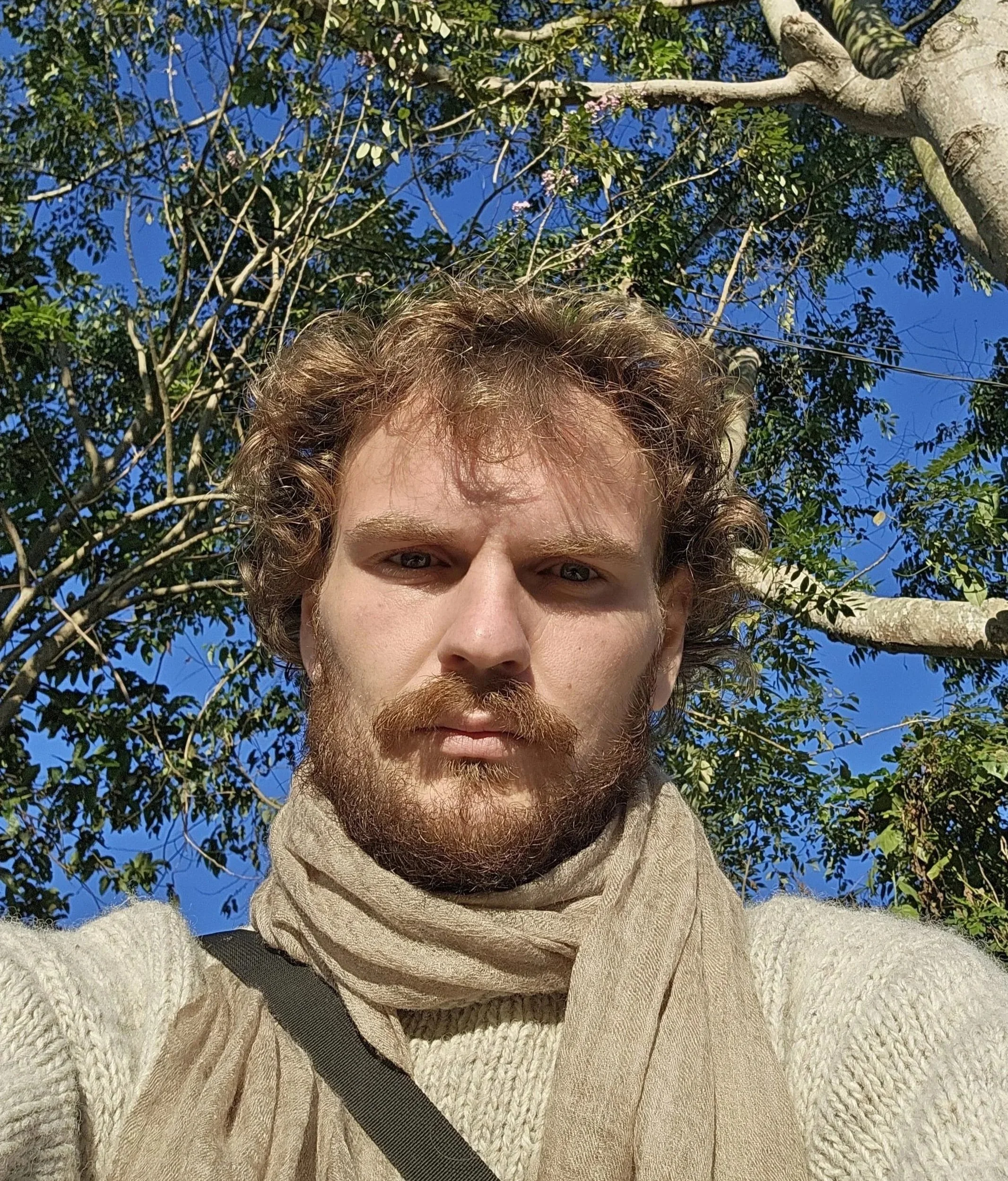 Een close-up van een man met roodbruin, golvend haar en een baard, gekleed in een lichtkleurige, gebreide trui en een beige sjaal, met een boom en blauwe lucht op de achtergrond.