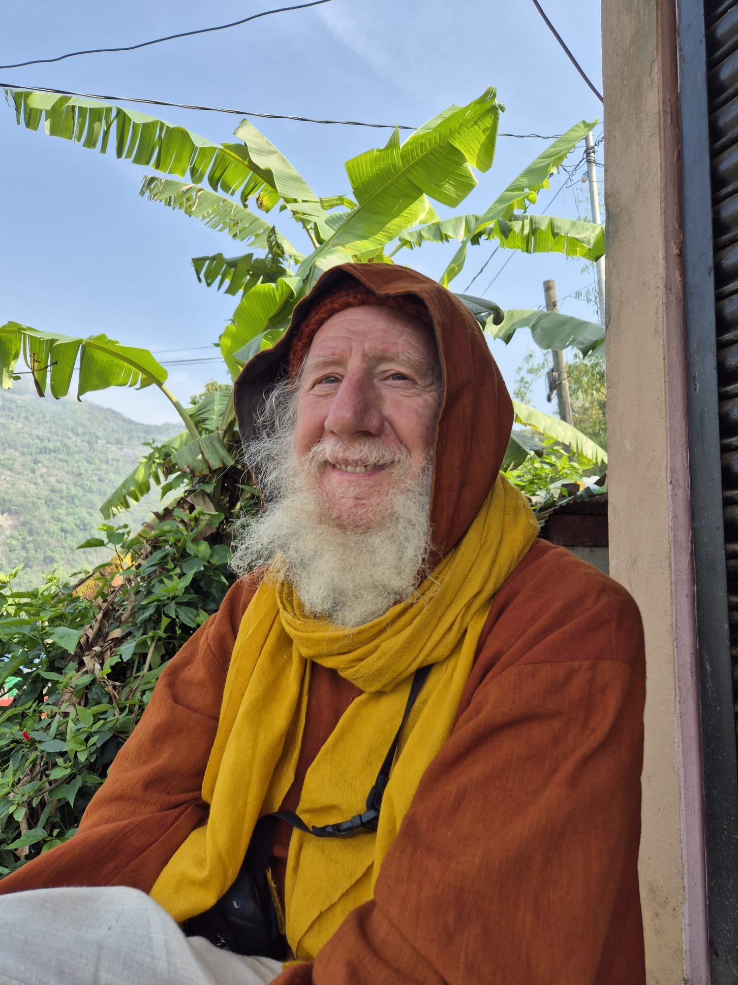 Een oude man met een grijze baard draagt een oranje hoodie met een kap en een gele sjaal. Hij is buiten, met een achtergrond van banana-bomen, een blauwe lucht en een heuvel met begroeiing.