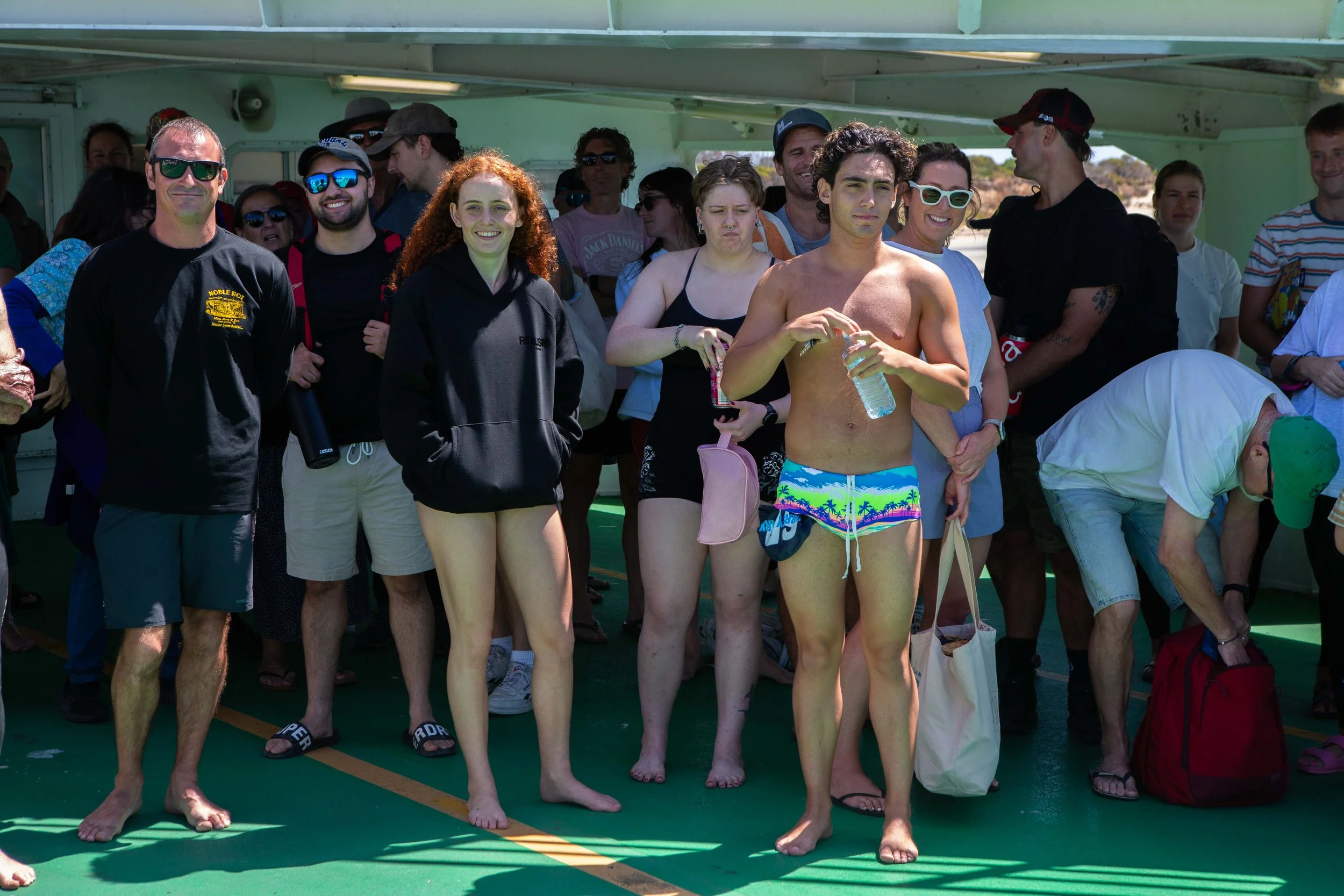 Group of people on a boat deck, some wearing swimsuits and casual clothes, smiling and relaxing, with a sunny outdoor background.