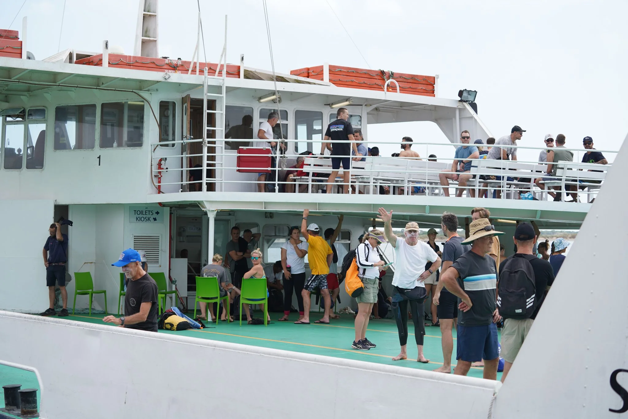 People standing and sitting on the deck of a ferry or cruise ship, some wearing hats and sunglasses, with a sign pointing to toilets and kiosk area visible on the lower level.