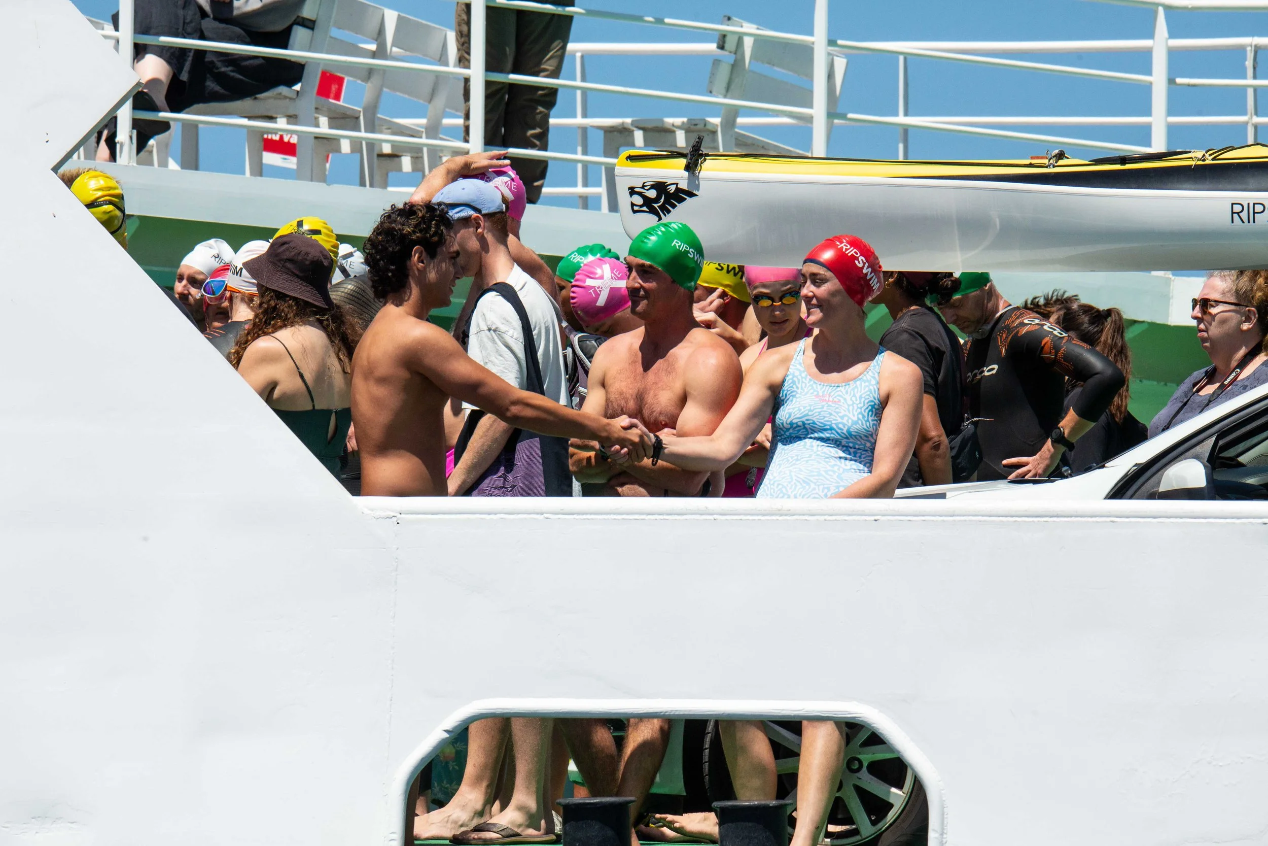 Swimmers in swim caps and goggles shaking hands on a boat deck after a race, with a rowing shell on the roof of the boat.