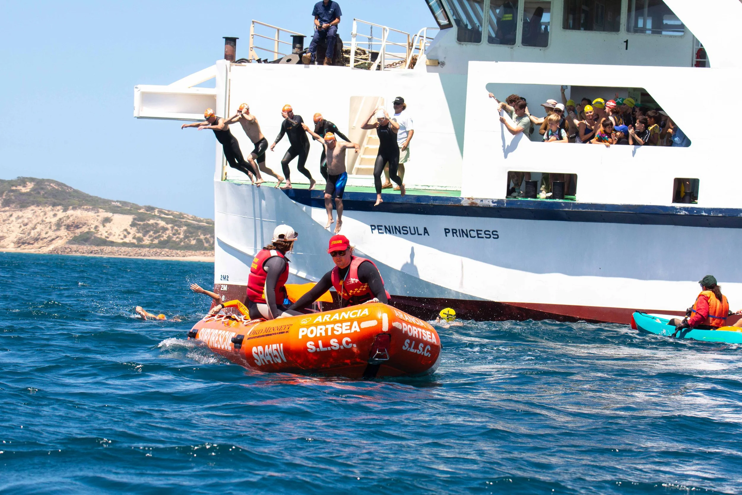 People jumping off a large white boat named 'PENINSULA PRINCESS' into the water, some rescuers in orange life vests nearby, with a hilly coastline in the background on a sunny day.