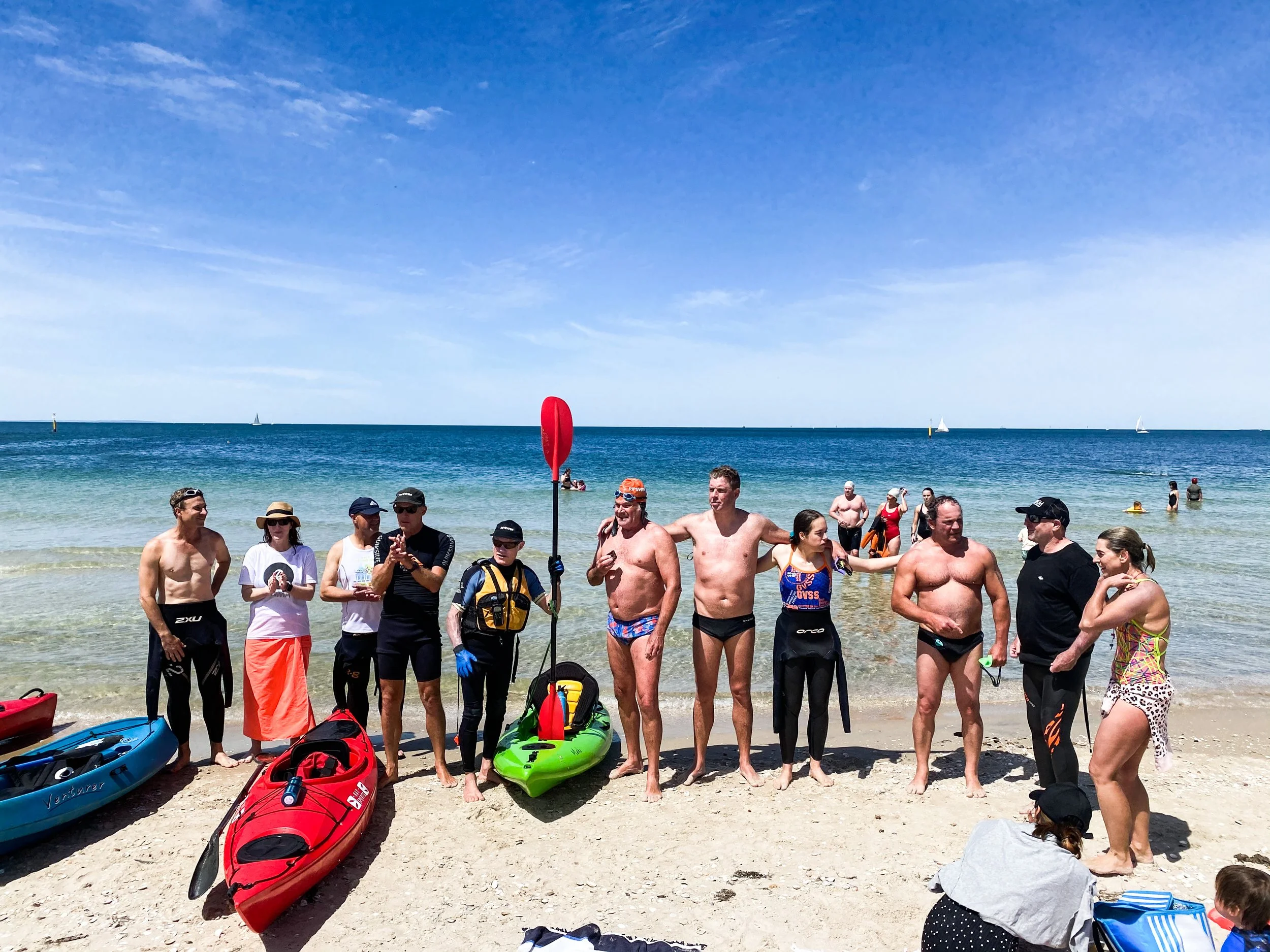 Group of people at the beach, some holding kayak paddles, with kayaks on the sand, and sailboats in the water under a blue sky.