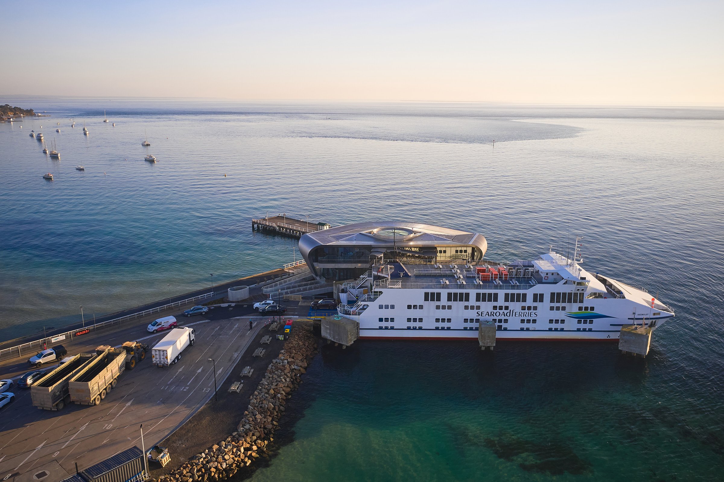 A ferry docked at a harbor with several boats floating in calm water. The background shows a clear sky and distant shoreline.