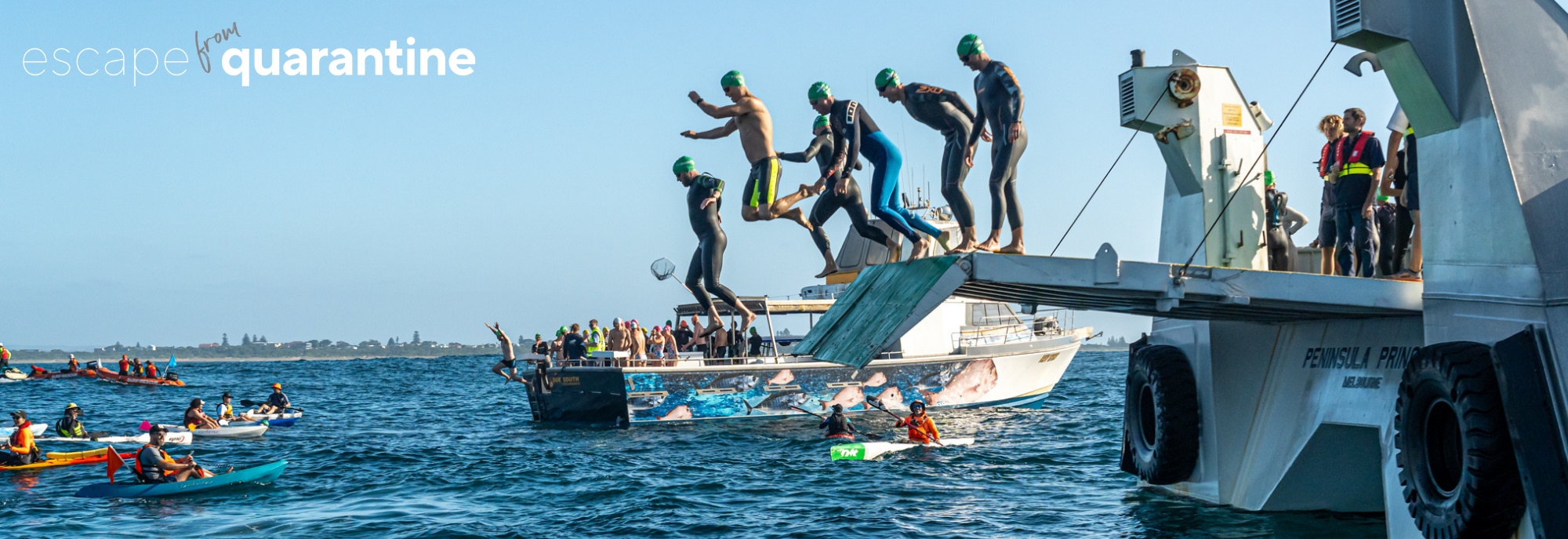 Swimmers jumping off a pier into the water during a swimming event, surrounded by kayakers and safety boats, with a backdrop of a coastal town.