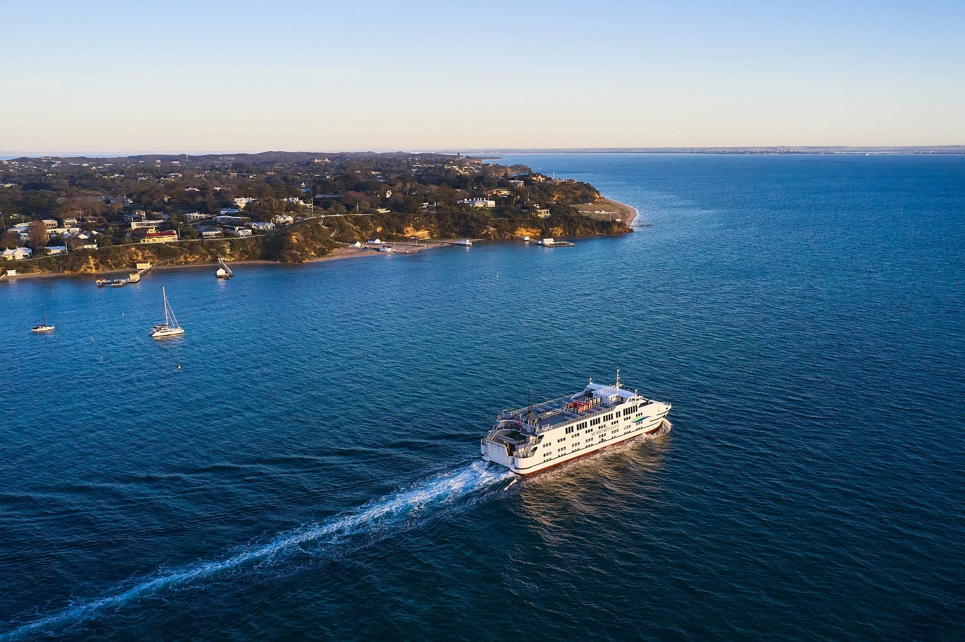 Aerial view of a coastal area with a large white ferry boat sailing in the blue water, other small boats anchored near the shoreline, and houses along the hillside overlooking the water.