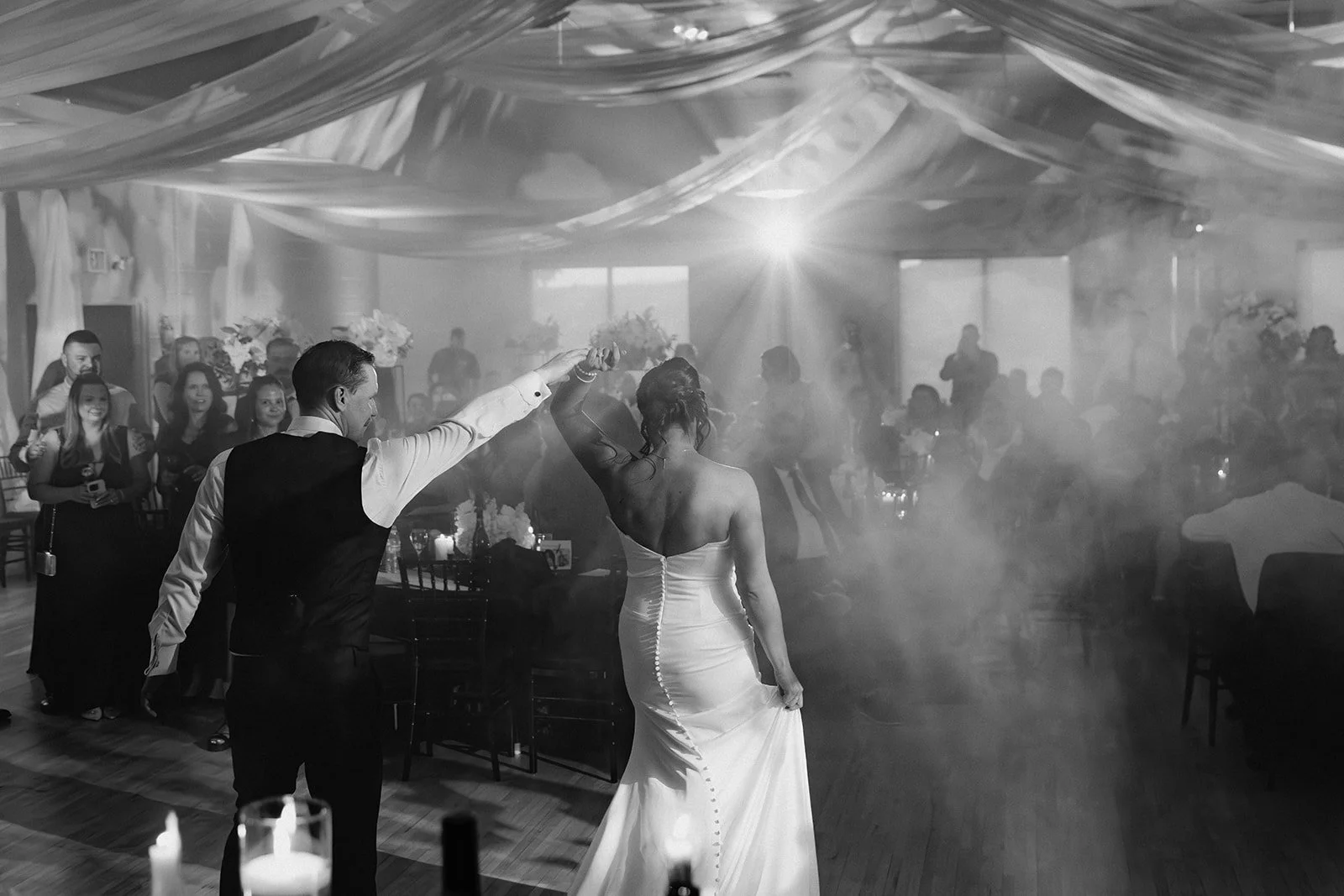 A bride and groom are dancing at their wedding reception, surrounded by guests and decorated with draped fabric and floral arrangements, in black and white.
